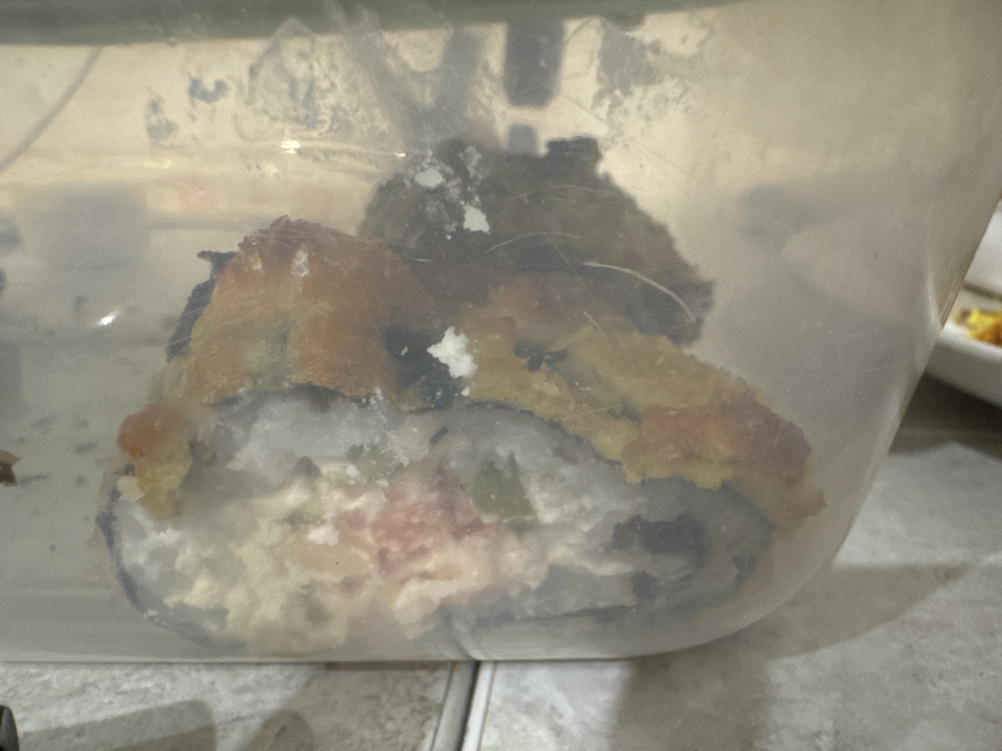 A close-up, eye-level, medium shot shows a transparent plastic container holding a deep-fried sushi roll that has been cut into several pieces. The sushi roll appears to be a California roll, with visible ingredients including rice, avocado, imitation crab, and possibly cream cheese. The exterior of the roll is coated in a golden-brown tempura batter, some of which has flaked off. The container is resting on a textured, light-colored surface, possibly a countertop or table, with faint tile lines visible. The background is out of focus, showing a light-colored wall with indistinct shadows and light reflections. There are no people present in the image. The scene is indoors, and no specific location within Saratov, Russia can be determined from the visual information provided.