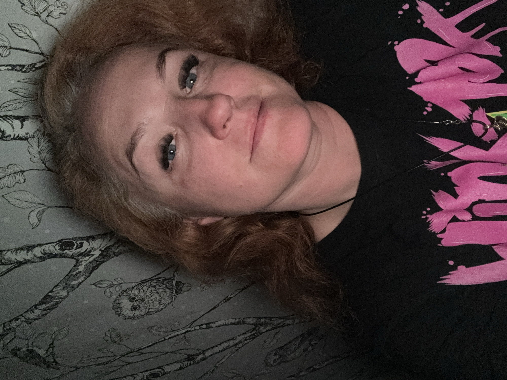 A woman with light skin and reddish-brown hair is lying on a surface with a muted green and grey pattern resembling a tree with an owl. She is wearing a black t-shirt with pink graphic elements. She has blue eyes and is looking at the camera with a slight smile. A thin black necklace is visible around her neck. There's no indication of the location being Chaikovsky, Russia.