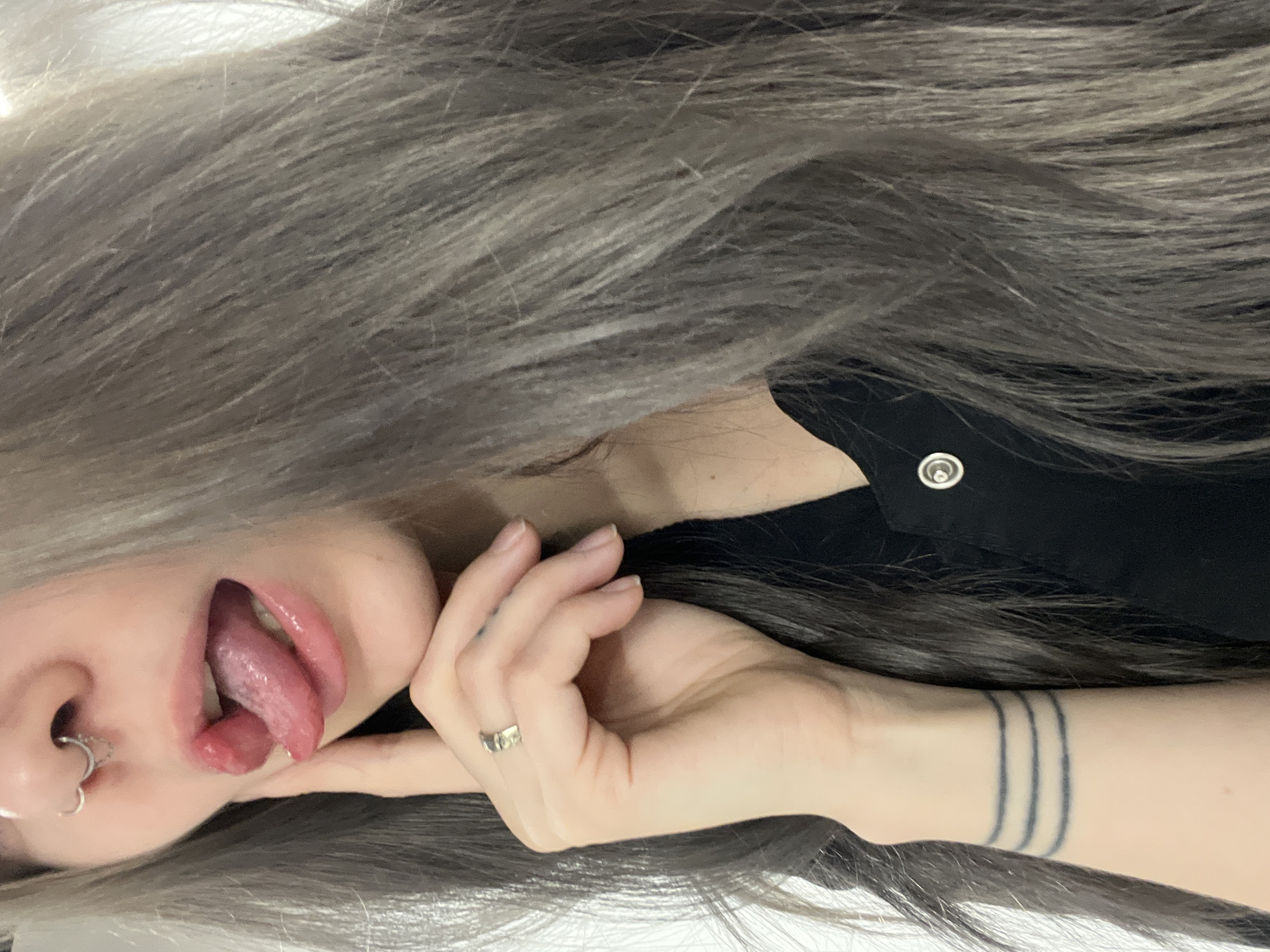 A close-up shot of a person with gray hair. The person's face is partially visible, with a focus on the mouth and tongue sticking out. A nose piercing is visible. The person's hand is positioned near their face and displays a ring. The wrist is visible and features a tattoo of three parallel lines. The person is wearing a black shirt or garment with a button.
The location in the city of Khimki, Russia, cannot be determined from the image.