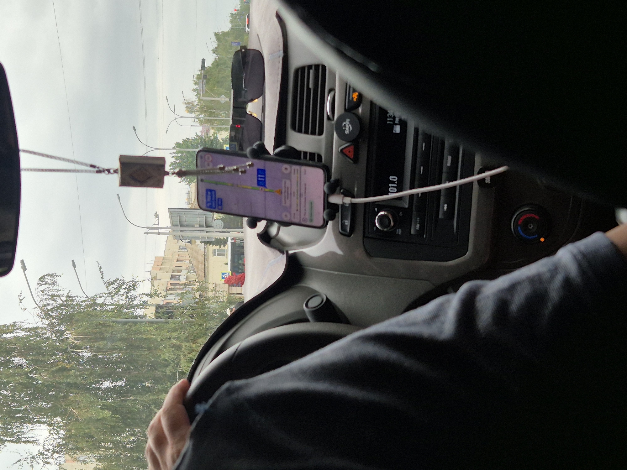 A driver's left hand grips the steering wheel of a car as they navigate through Kokshetau, Kazakhstan. The car's dashboard is visible, featuring a smartphone mounted on a holder, displaying navigation. A beaded car charm hangs from the rearview mirror. Outside the car, buildings and trees line the street, under a cloudy sky. The scene suggests an urban setting, possibly a residential area given the presence of apartment buildings and streetlights. The overall atmosphere is one of everyday travel.