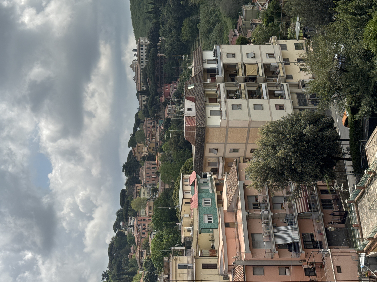 This vertical image, when correctly oriented, showcases a dense hillside town, characteristic of Ariccia, Italy, under a heavily overcast sky. Numerous multi-story residential buildings, varying in color from pale yellow and cream to light pink and green, are nestled across a steep, tree-covered slope. Terracotta-tiled roofs top many of the structures.
Abundant green trees and dense foliage are interspersed throughout the town, particularly along the upper sections of the hill, creating a harmonious blend of urban and natural environments. In the distance, on the highest point of the hill, a grander, lighter-colored building with a distinct dome or prominent roof structure is visible, hinting at a significant historical or public building.
A winding road can be seen in the mid-ground, where a single, light-colored car is parked. Many of the buildings feature balconies, some equipped with striped awnings or visible laundry, indicating active residential use. The sky is dominated by large, grey clouds, suggesting a cloudy day. No specific human activity is discernable, and there is no visible text.