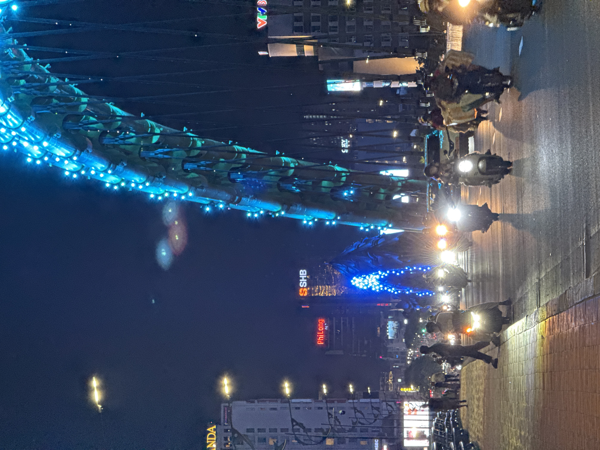 This night-time photograph captures a vibrant cityscape in Đà Nẵng, Vietnam, centered around an illuminated bridge and a busy street. The scene is bathed in artificial light against a dark blue sky.
On the left side, a prominent bridge arches across, its structure adorned with numerous bright blue LED lights, suggesting a festive or ornamental illumination. The bridge's design features intricate, scale-like panels, reminiscent of a dragon, a characteristic feature of the iconic Dragon Bridge in Đà Nẵng. The blue lights highlight the bridge's curvature and texture.
To the right, a street is bustling with activity. Several motorbikes, their headlights and taillights glowing, indicate ongoing traffic. One motorbike with a bright headlight is clearly visible in the mid-ground. Pedestrians are also present on the sidewalk to the far right, suggesting evening strolls or commutes. The street surface reflects some of the surrounding lights, indicating a possibly damp or smooth surface.
In the background, behind the street and bridge, numerous multi-story buildings rise into the night sky. Many of these buildings have illuminated windows and vibrant neon signs. Visible text includes "SHB" in orange and white, and "PhiLong" in red on one of the taller buildings. Other partial text like "VIVA" and "ANDA" are also discernable on different structures.
The image captures the lively atmosphere of the city at night, with the illuminated bridge serving as a striking focal point. The bright lights from vehicles, buildings, and the bridge itself create a dynamic and energetic urban environment.