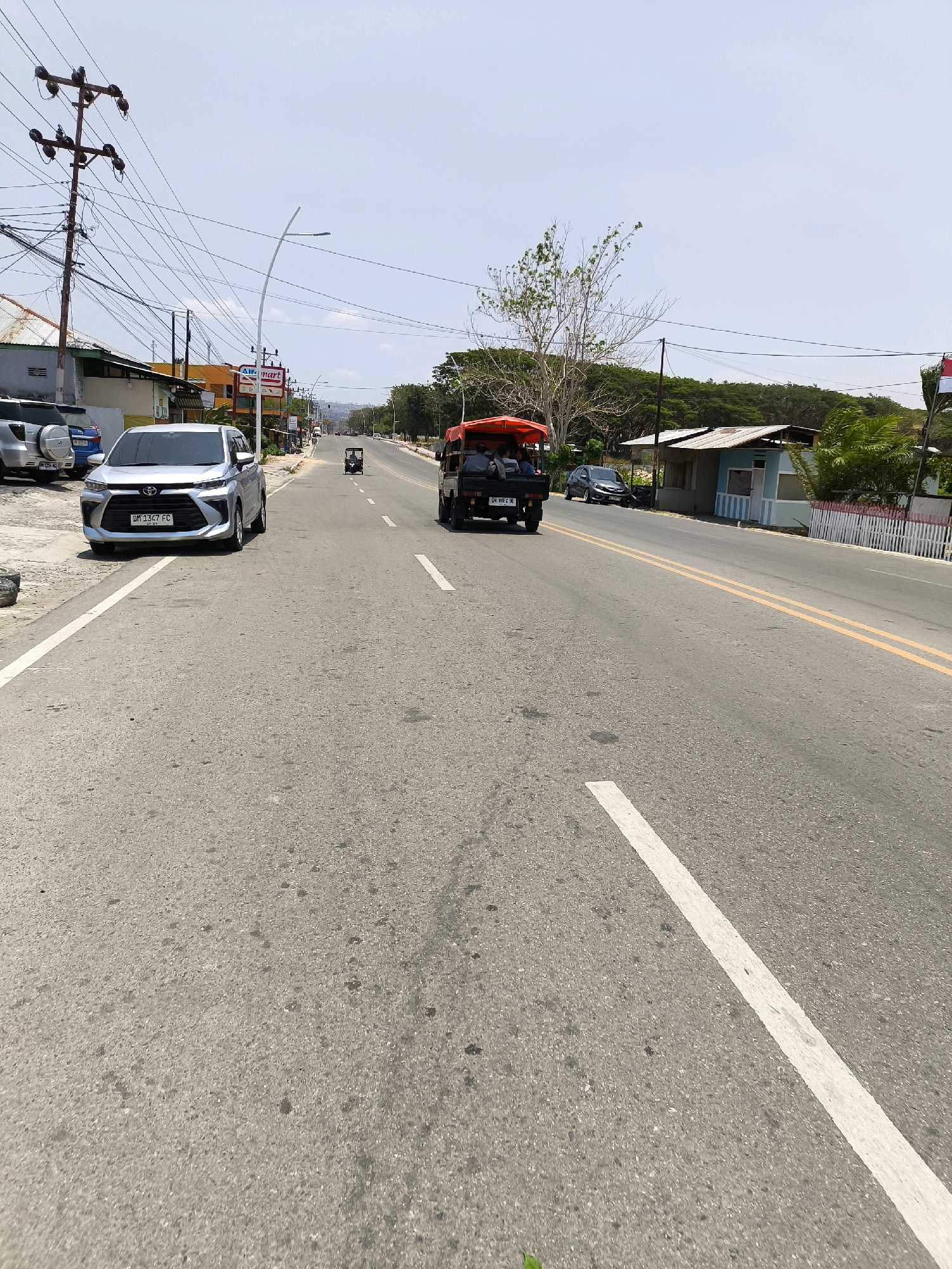 The image depicts a street scene in Kwandang, Indonesia, on a clear, sunny day. A paved road with white and double yellow lines runs towards the horizon. On the left side of the road, a silver Toyota Avanza is parked, with other vehicles and buildings visible in the background, including a Mart convenience store. On the right side of the road, a small, open-air vehicle, possibly a type of tuk-tuk or small utility vehicle, is driving away from the camera, carrying passengers. Further down the road, another similar vehicle is visible. Trees and vegetation line the sides of the road, with some houses and structures also present. Electrical poles and wires are prominent on the left side of the street. The overall impression is of a typical, active street in a coastal or tropical urban environment.