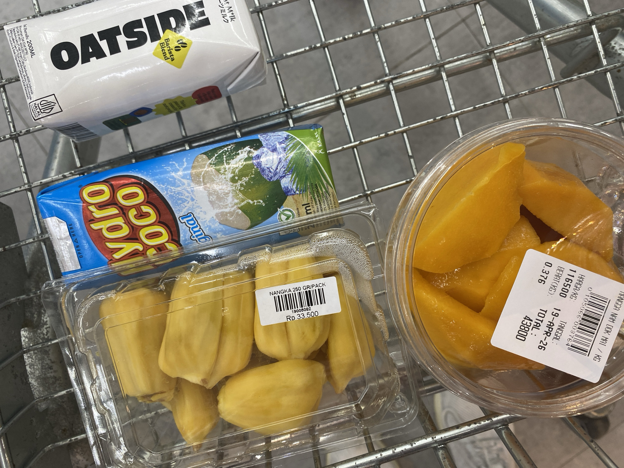 The image depicts a selection of grocery items in a metal shopping cart. The items include a carton of OATSIDE Barista Blend oat milk, a carton of Hydro Coco Original coconut water, a clear plastic container of jackfruit pieces labeled "NANGKA 250 GR/PACK Rp 33.500", and a clear plastic container of sliced mango pieces labeled with price and weight information, "MANGGA NAM DOK MAI / KG", "116500", "19-APR-26", "0.376", "43800". Based on the food products' labels (PT ABC Kogen Dairy, Bandung and PT Pulau Sambu Riau 29255) and the use of Indonesian Rupiah (Rp), the location is assumed to be Indonesia. No people are visible. The image does not confirm it is Kuta Selatan.