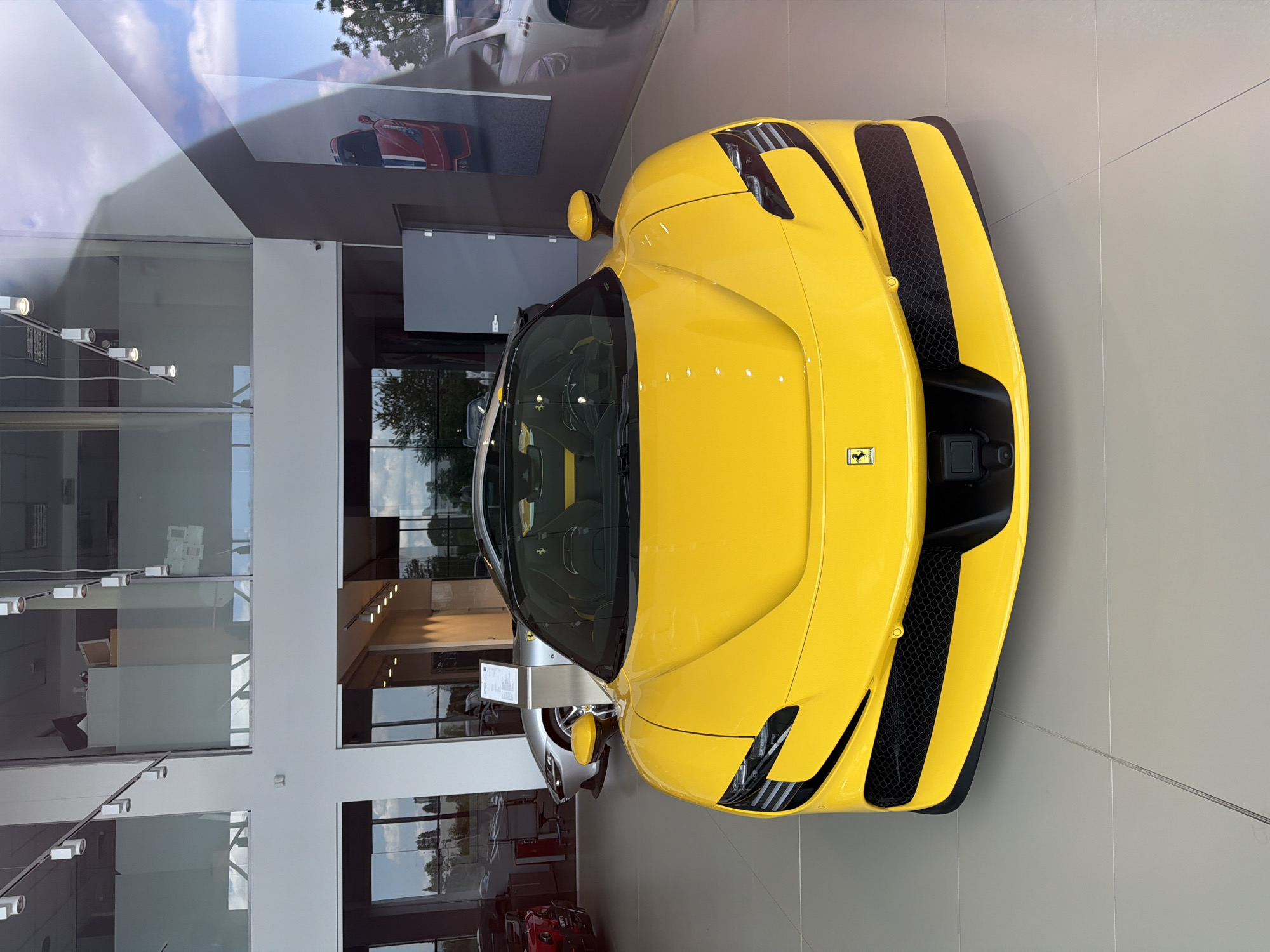 A bright yellow Ferrari sports car is the central subject of this image, prominently displayed indoors within what appears to be a modern car dealership or showroom in Busmantsi, Bulgaria. The car is oriented with its front facing towards the bottom right of the frame, showcasing its sleek design, distinctive headlights, and black grille accents. A small Ferrari emblem is visible on its front hood.
The car is stationary, positioned on a light-colored tiled floor. Behind the yellow Ferrari, another silver or grey sports car is partially visible, also on display, with an unreadable information stand placed beside it.
The setting is well-lit, suggesting daytime. Large glass walls and windows enclose the showroom, reflecting the exterior environment. Reflections of white clouds and green trees are visible on the glass and the car's glossy yellow surface, indicating clear weather outside. Posters or large screens on the showroom walls depict other cars, including a red sports car. The interior features modern architecture with white pillars, ceiling lights, and minimalist design. A small security camera is mounted on one of the white pillars.
No people are visible in the image, emphasizing the car and the showroom's pristine presentation. The scene conveys a sense of luxury and high-end automotive display.