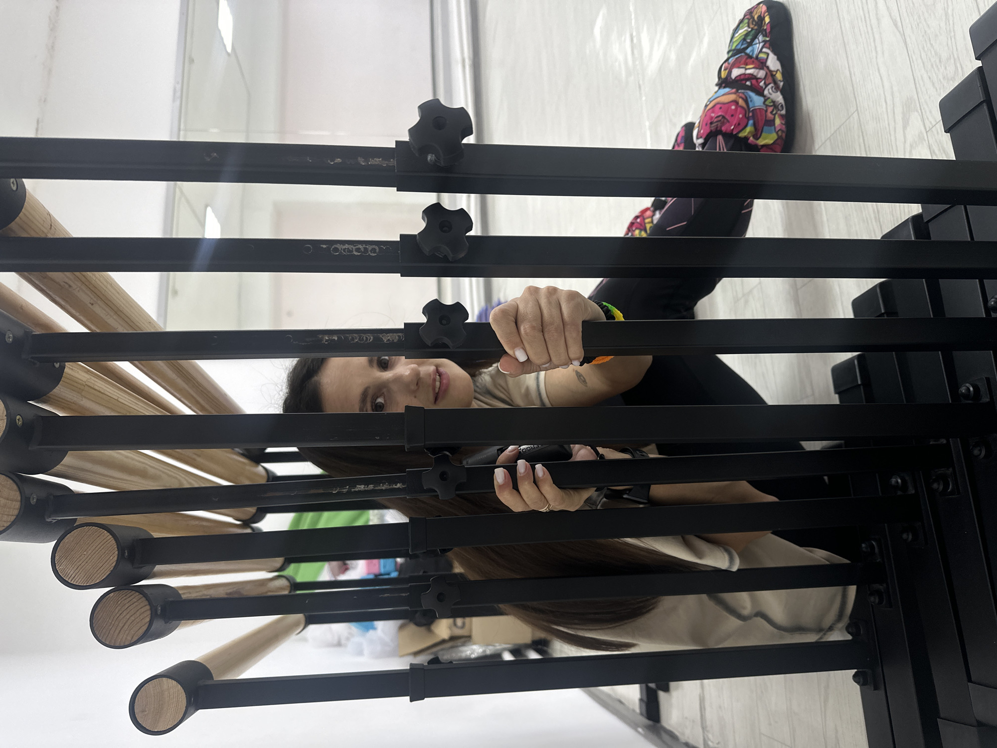 The image, rotated 90 degrees counter-clockwise, captures a woman peeking through a black metal framed structure, likely a multi-tiered exercise or dance barre. She has long dark hair and is looking directly at the viewer with a slight smile, appearing friendly and relaxed. She wears a light-colored top and dark leggings, with a vibrant, multi-colored patterned slipper or shoe visible on her left foot in the upper right corner. A colorful bracelet adorns her left wrist.
Her left hand grips one of the black horizontal bars of the structure, while her right hand holds a black object, possibly a phone or camera, also through the bars. The structure itself is composed of black metal frames with several wooden cylindrical bars inserted horizontally, featuring black star-shaped knobs for adjustment.
The setting appears to be a bright, indoor space in Votkinsk, Russia, with white walls and a light-colored, possibly wooden or laminate, floor. The overall impression is clean and well-lit, typical of a studio, gym, or a modern room. The woman seems to be posing for the camera, perhaps taking a break from an activity or capturing a casual moment.
Visible text includes faint lettering on one of the black metal bars, possibly "OOOFO," and "ERGO" is partially visible on the object held in her right hand. The time of day is not evident, but the good interior lighting suggests daytime.
