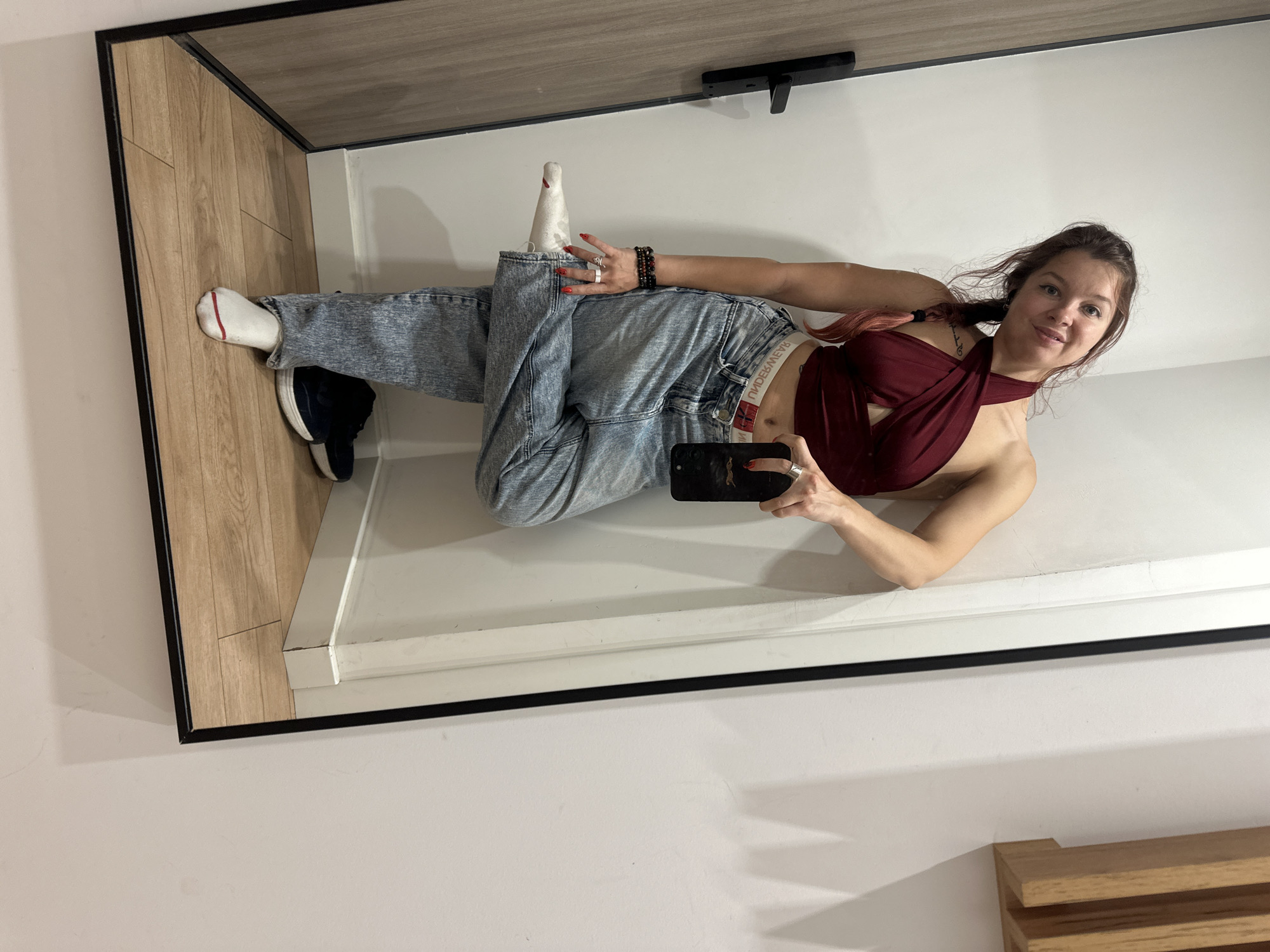 The image is a mirror selfie of a young woman. She is lying down on a floor with a wood-patterned surface. She is wearing a maroon halter top, light-wash denim jeans, white socks, and dark sneakers. She has on multiple rings and bracelets. The woman is holding a smartphone in her right hand, which she is using to take the picture. Her hair is dark brown with pink highlights and is pulled back into a ponytail. Her expression is a slight smile.
The background consists of a white wall and a large mirror that reflects the scene. The mirror is framed in black. A light-colored shelf or ledge is visible below the mirror, and a dark, rectangular object is mounted on the wall above the ledge. The lighting suggests it is daytime, possibly indoors with artificial lighting. The overall setting appears to be a domestic environment, perhaps a bedroom or dressing area in Moscow, Russia. The woman's pose and the casual attire suggest a relaxed and informal moment.
