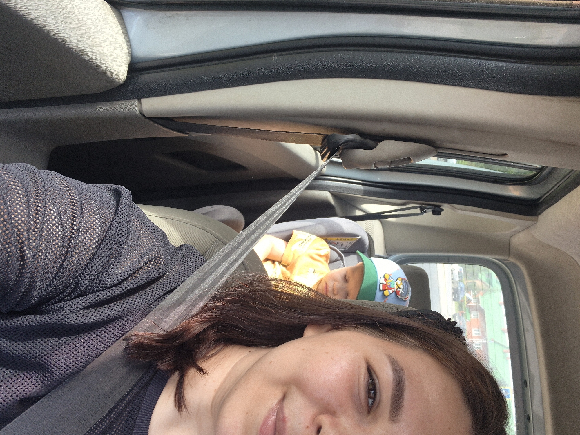 In a vehicle, a woman is taking a selfie while a young child sits in a car seat behind her. The woman is wearing a black mesh shirt and a seatbelt. The child is wearing an orange shirt and a blue and green cap with a cartoon character on it. The vehicle's interior is visible, including the door panel, seat, and a portion of the rear window showing houses and trees outside. The scene appears to be on a street in Chaikovsky, Russia.