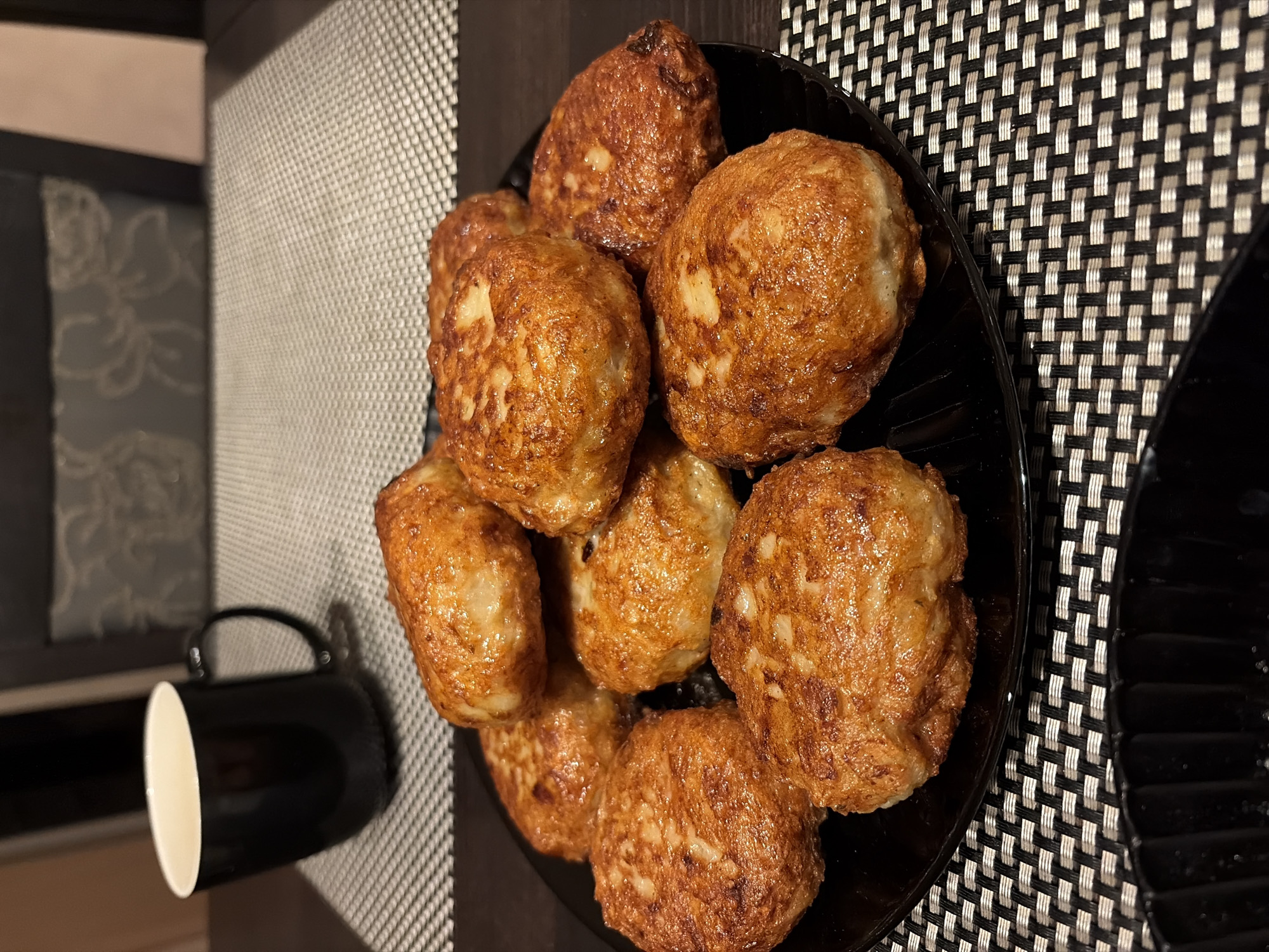 A black plate piled with golden-brown, spherical food items, possibly fried meatballs or croquettes, sits on a patterned placemat of alternating black and white squares. The placemat is placed on a dark-colored surface. To the left of the plate is a black coffee mug with a white interior. The background includes a dark-colored wall or furniture. The food items appear to be freshly cooked and neatly arranged on the plate. The lighting suggests an indoor setting, potentially a restaurant or dining room. There is no indication that the photo was taken in Budva, Montenegro. The image does not contain people or actions.