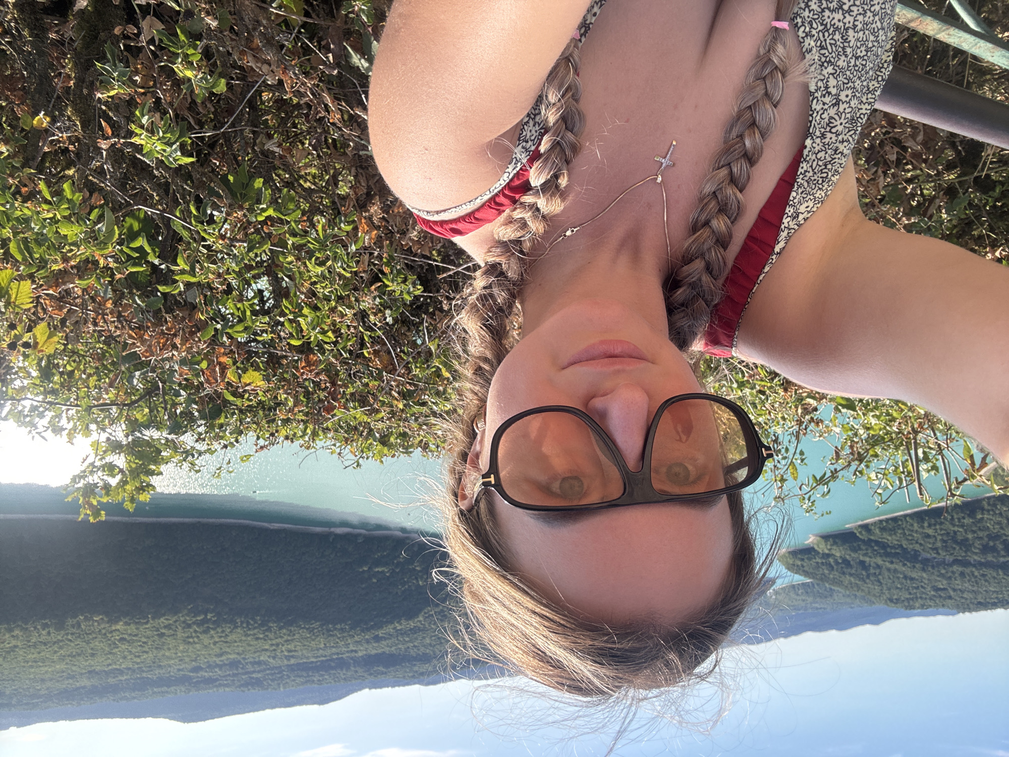 A woman is taking a selfie on a sunny day, with a large turquoise body of water and forested hills in the background. The woman has blonde hair in two braids and is wearing large, dark-rimmed sunglasses. She is also wearing a dress with a floral pattern and a cross necklace. The scene appears to be a scenic overlook, possibly a viewpoint or a park, in or near Moirans-en-Montagne, France, given the mountainous terrain and body of water. The vegetation in the foreground consists of bushes and trees.