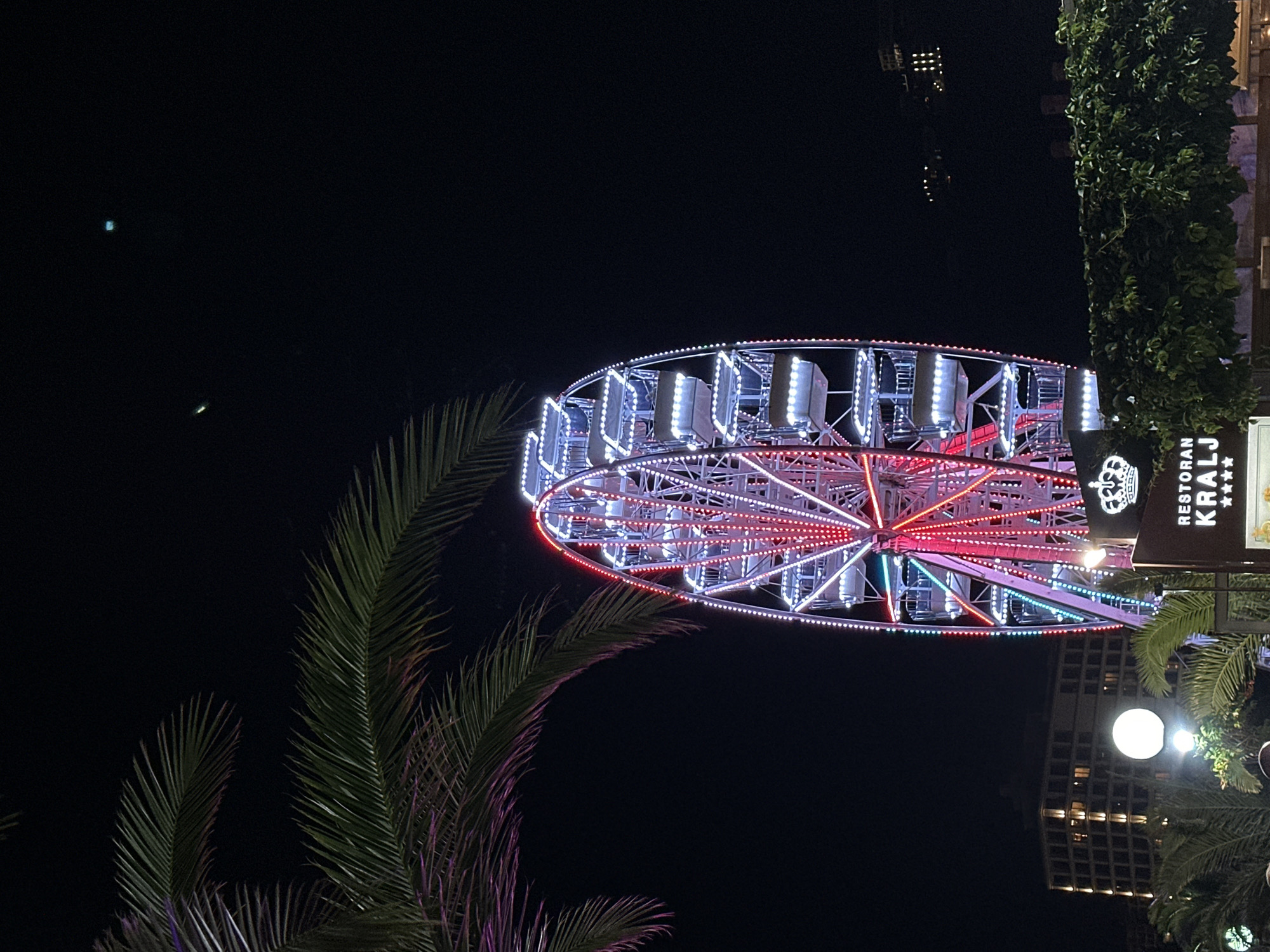 An exterior night view captures a Ferris wheel illuminated with vibrant red and white lights in Budva, Montenegro. The Ferris wheel stands tall against a dark sky, its spokes and cabins clearly defined by the colorful illumination. In the foreground, a palm tree's fronds are visible, partially obscuring the sky and framing the upper left portion of the Ferris wheel. To the left of the Ferris wheel, a multi-story building with illuminated windows is visible, suggesting an urban setting. In the lower foreground, a sign for "RESTORAN KRALJ" is partially visible, adorned with a crown emblem and stars. Lush green foliage partially obscures the base of the scene. The overall ambiance is festive and recreational.