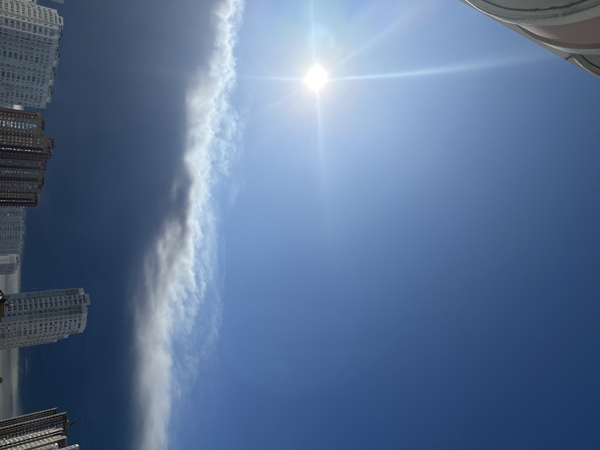 The image shows a dramatic sky over the city of Podolsk, Russia. The sun is shining brightly, creating a lens flare in the upper left quadrant of the image. Below the sun, a distinct band of dark, ominous clouds stretches across the sky, contrasting sharply with the clear blue sky above. A lighter, wispy layer of clouds is visible just above the dark band. In the lower portion of the image, several multi-story buildings are visible, their windows reflecting the sky. The perspective is from a low angle, looking up at the sky and the tops of the buildings. There are no people or specific actions depicted in the image. The environment appears to be urban, with the buildings suggesting a residential or commercial area.