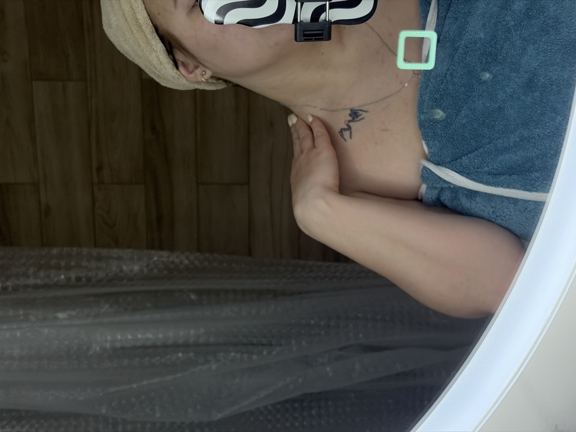A person is visible in the image, likely reflected in a mirror. The person has a towel wrapped around their head and is wearing a blue garment. They have a tattoo on their chest/neck area and are wearing a necklace. The background includes what appears to be a wood-paneled wall and some type of plastic-wrapped material. There's an object attached to their head with black and white patterns. The location is unspecified, with no clear evidence linking it to Krasnodar, Russia.