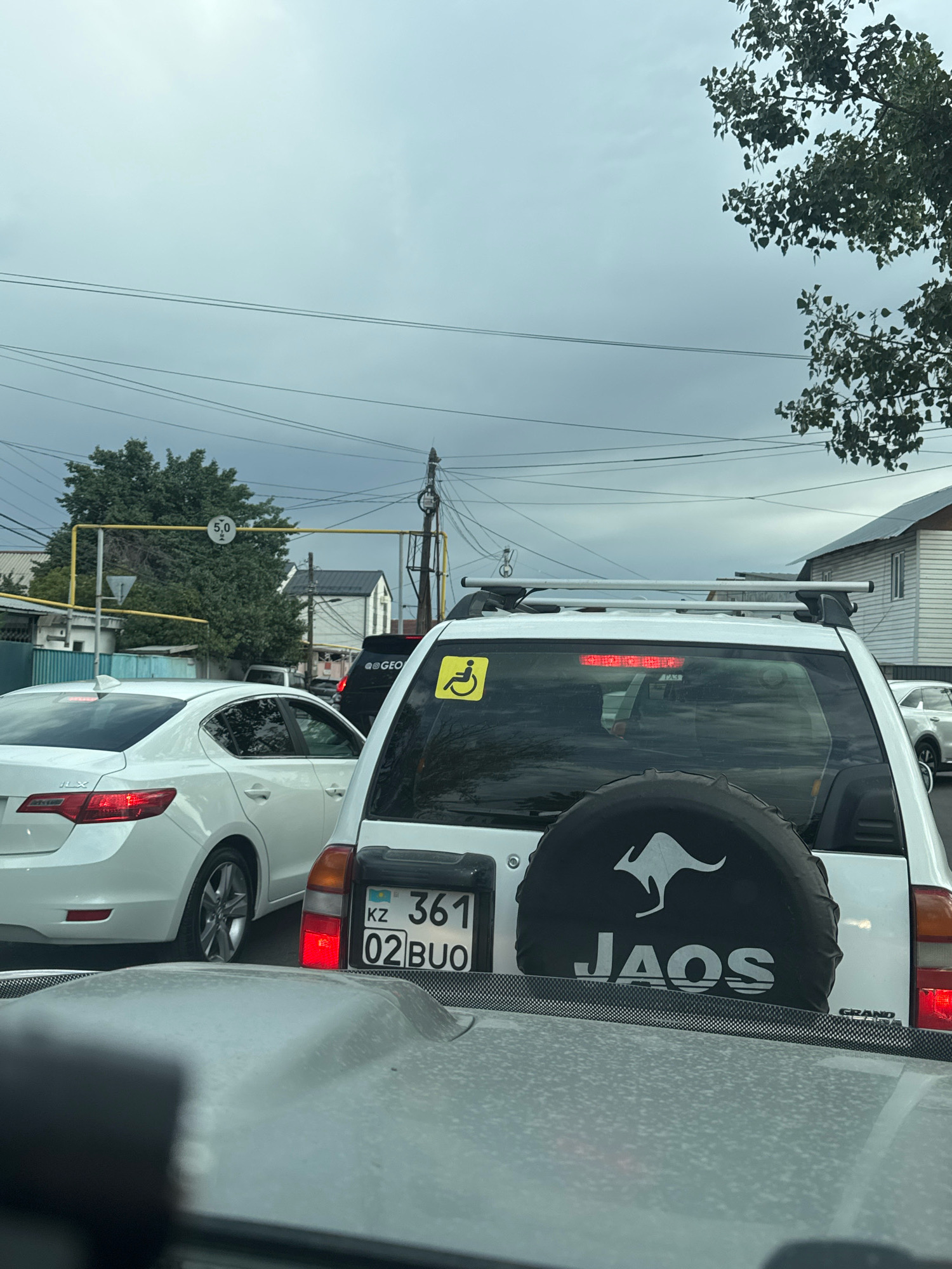 In Almaty, Kazakhstan, on an overcast day, a traffic jam is visible. The foreground shows the hood of a car, reflecting the sky. Behind it, a white Suzuki Grand Vitara is prominent, featuring a spare tire cover with a white kangaroo silhouette and the text "JAOS". A yellow wheelchair accessibility sticker is affixed to its rear window. The vehicle's license plate reads "KZ 361 02 BUO". To the left, a white sedan is partially visible. A yellow sign indicating a speed limit of 5.0 is suspended over the road, accompanied by an overhead power line. In the background, houses and trees can be seen.