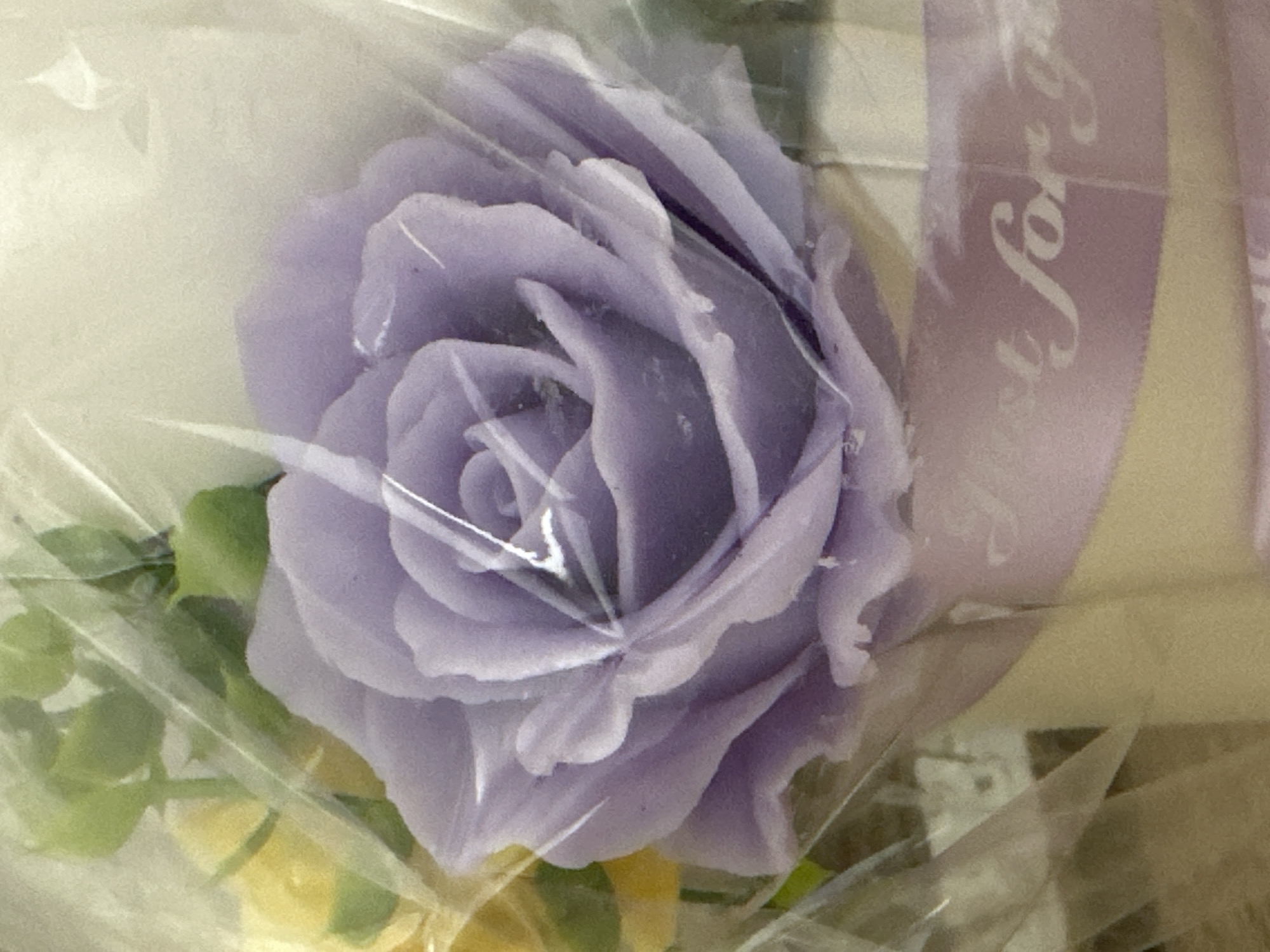 The image shows a close-up view of a lavender rose, likely a candle or a decorative element, wrapped in clear plastic. The rose is detailed with layered petals and is positioned in the foreground. To the left and slightly behind the rose, there are hints of yellow flowers and green foliage, also appearing to be artificial. A light purple ribbon with white text, partially obscured by the plastic and the rose, is visible across the bottom portion of the image. The text on the ribbon appears to read "Just for you". The background is a soft, out-of-focus light color. The image is a still life and does not depict any specific location in Yessentuki, Russia, nor does it contain any people or actions.