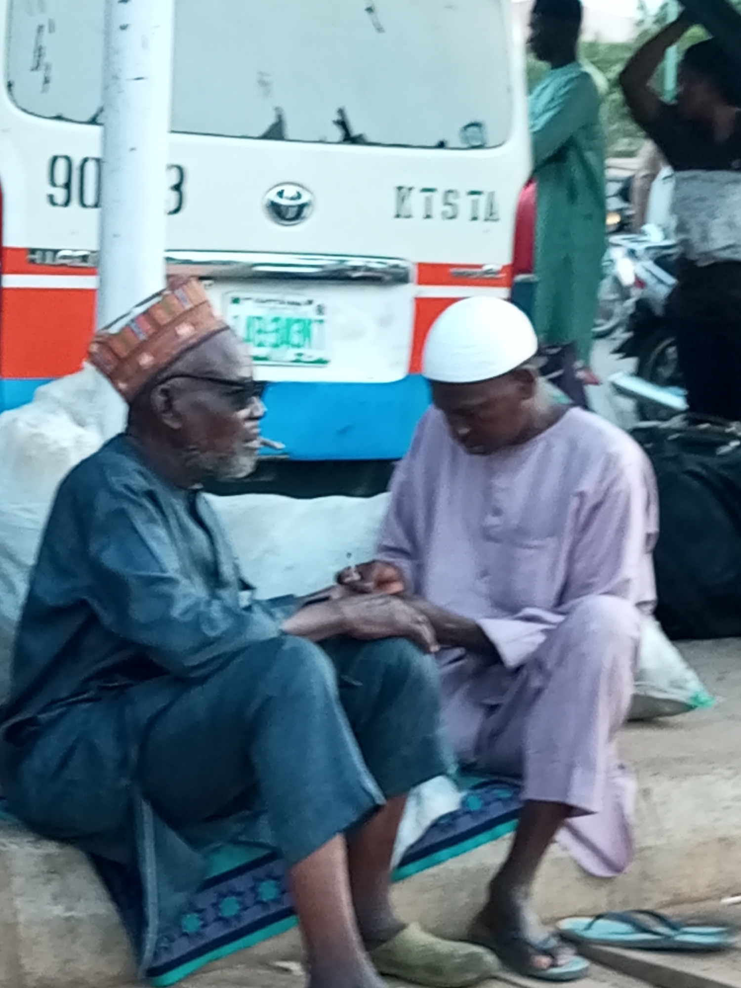 The image depicts two men sitting on a curb in what appears to be a busy urban area. The man on the left is wearing a dark blue outfit and a patterned cap, with glasses and a beard. He is holding a cigarette. The man on the right is wearing a light purple outfit and a white skullcap. They are seated next to a white bus with the numbers "903" visible on its side, and a license plate with green text. The background suggests a bustling environment, possibly a bus station or market, with other people and vehicles partially visible. The setting is consistent with the environment of the city of Gombe, Nigeria.