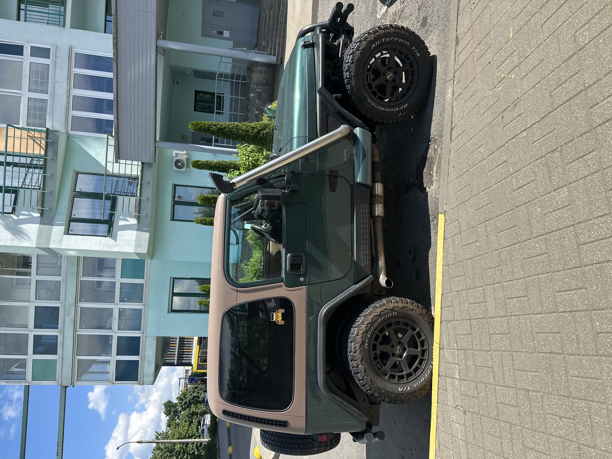 This is a forensic, matter-of-fact description of the image:
The image shows the side profile of a dark green Jeep Wrangler parked on a brick-paved sidewalk. The Jeep has a tan hardtop and a snorkel intake. It is equipped with off-road tires and black rims. A spare tire is mounted on the rear of the vehicle. In the background, there is a light blue, multi-story building with numerous windows and balconies. To the left of the Jeep, a yellow line marks the edge of the sidewalk. Further in the background, trees and a yellow bus are visible. An air conditioning unit is mounted on the side of the building. The scene appears to be in a city environment, potentially Minsk, Belarus, based on typical architectural and street characteristics. No people are visible in the image. The lighting suggests it is daytime, with a clear sky and scattered clouds.