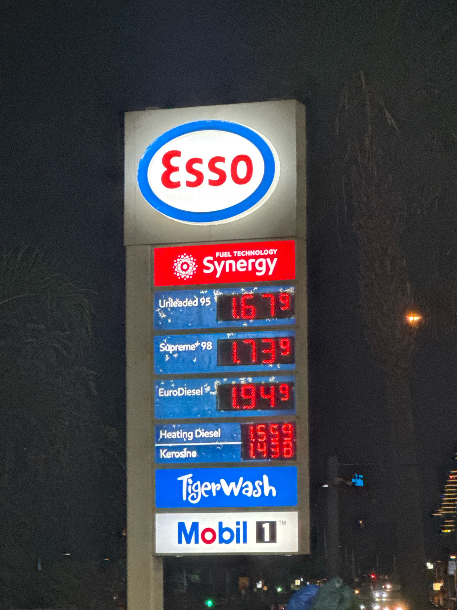 The image shows a nighttime view of a roadside Esso gas station sign in Agios Tychona Community, Cyprus. The sign displays fuel prices, with the Esso logo at the top. Below the logo, it reads "FUEL TECHNOLOGY SYNERGY" in red lettering against a red background. The prices are listed for "Unleaded 95" at 1.679, "Supreme+ 98" at 1.739, and "EuroDiesel" at 1.949. Further down, "Heating Diesel" is priced at 1.559 and "Kerosine" at 1.438. A blue sign reads "TigerWash," and below that, a white sign displays "Mobil 1™". The surrounding environment is dark, with some trees visible on the left and buildings and streetlights in the background on the right. It appears to be raining or snowing lightly, as indicated by small white specks in the air and on the sign.