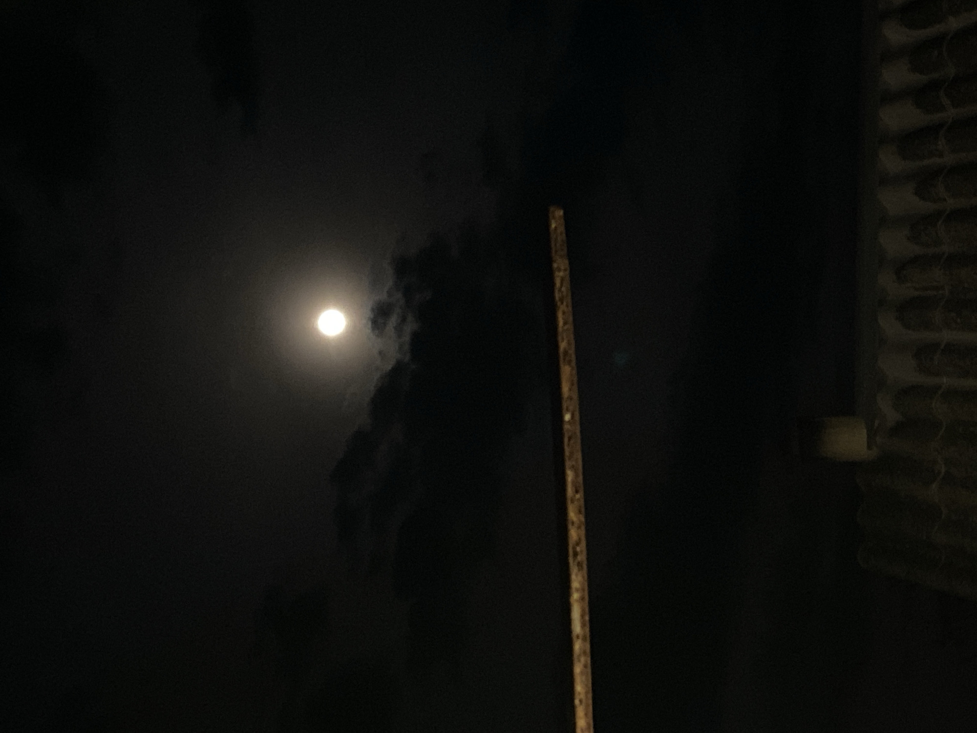 The image presents a nighttime scene featuring a bright, full moon partially obscured by dark, wispy clouds. The moon, positioned in the upper-left quadrant, casts a luminous glow that subtly highlights the contours of the surrounding clouds. A long, slender pole or stick, appearing weathered and possibly made of wood or bamboo, stands vertically in the right-center of the frame, extending from the bottom to the top. Along the far right edge of the image, a distinct textured structure is visible. It resembles a tiled roof or a corrugated surface, characterized by overlapping, wavy patterns. The sky is dark, indicating it is night, with significant cloud cover. No people or specific activities are depicted. The scene suggests an outdoor setting, potentially a view from a ground level or a balcony looking towards the sky and a nearby architectural element. No text is visible in the image.