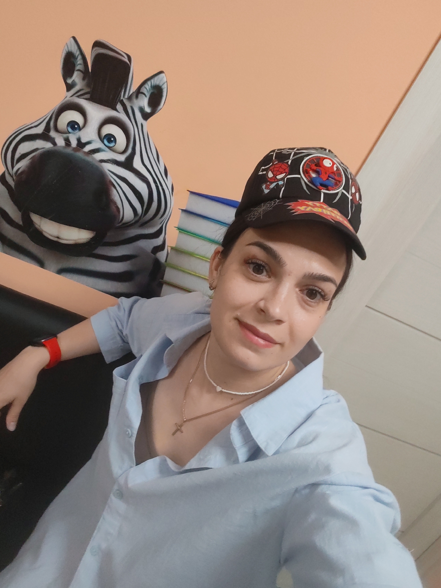 A young woman with dark hair, wearing a blue button-down shirt and a black baseball cap with Spiderman graphics, takes a selfie. She is wearing a white beaded necklace with a small cross pendant and a red fitness tracker on her left wrist. To her left, a large cartoon zebra decoration with wide blue eyes and an open mouth is visible against an orange wall. In the background, a stack of blue and white books is perched on a surface. The scene appears to be indoors, possibly in a room in the city of Belogorsk, Russia, given the context.