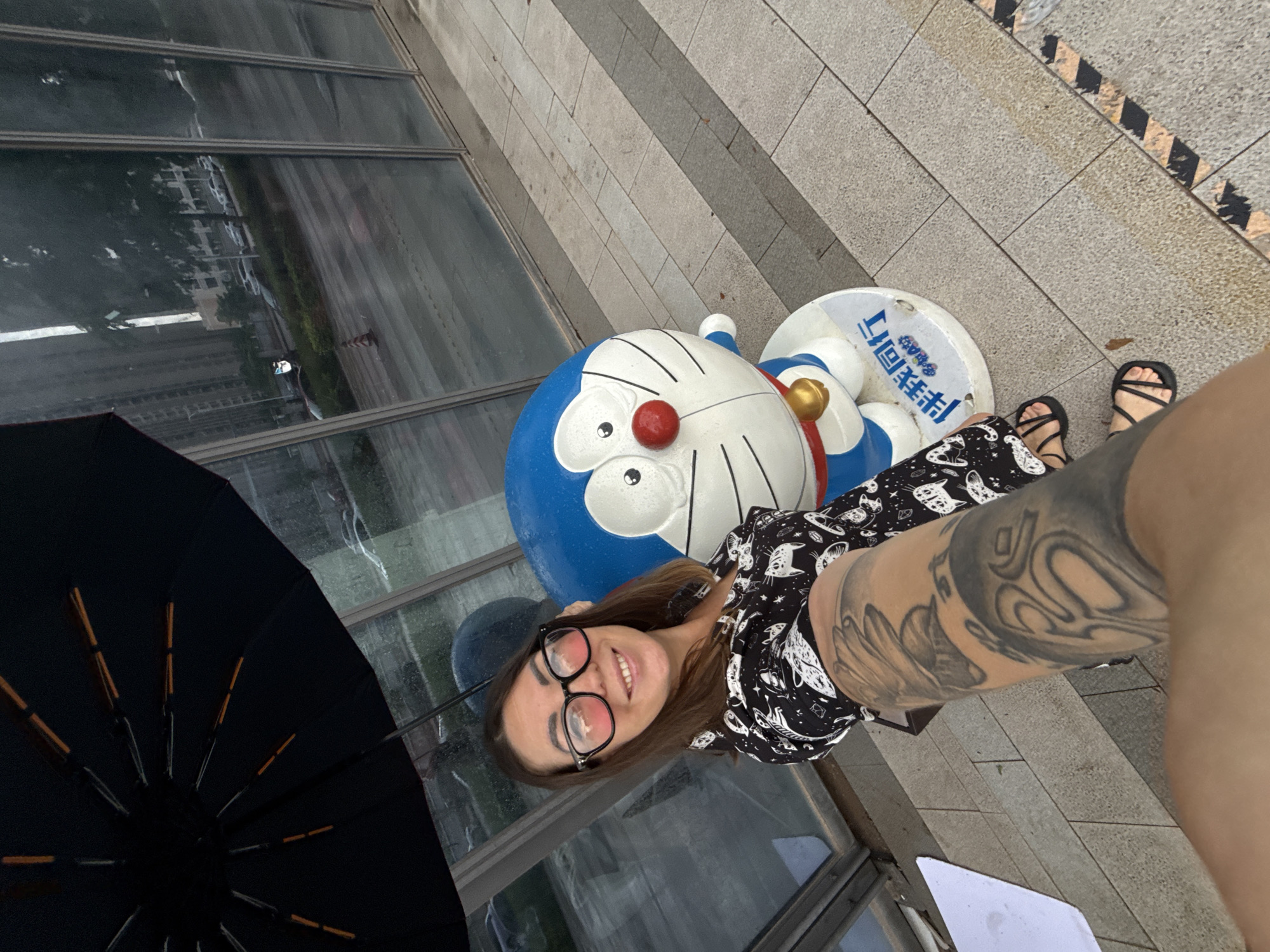 A woman is taking a selfie outdoors in Huizhou, China, next to a statue of Doraemon. She is holding an umbrella above herself, suggesting it is raining. The woman is wearing glasses, a black and white patterned shirt, and black sandals. She has extensive tattoos on her left arm. The Doraemon statue is blue and white with a red nose and a yellow bell. Behind them, large glass windows reflect a street scene with buildings and cars. The ground is paved with grey tiles. The overall impression is a casual moment captured on a potentially wet day.