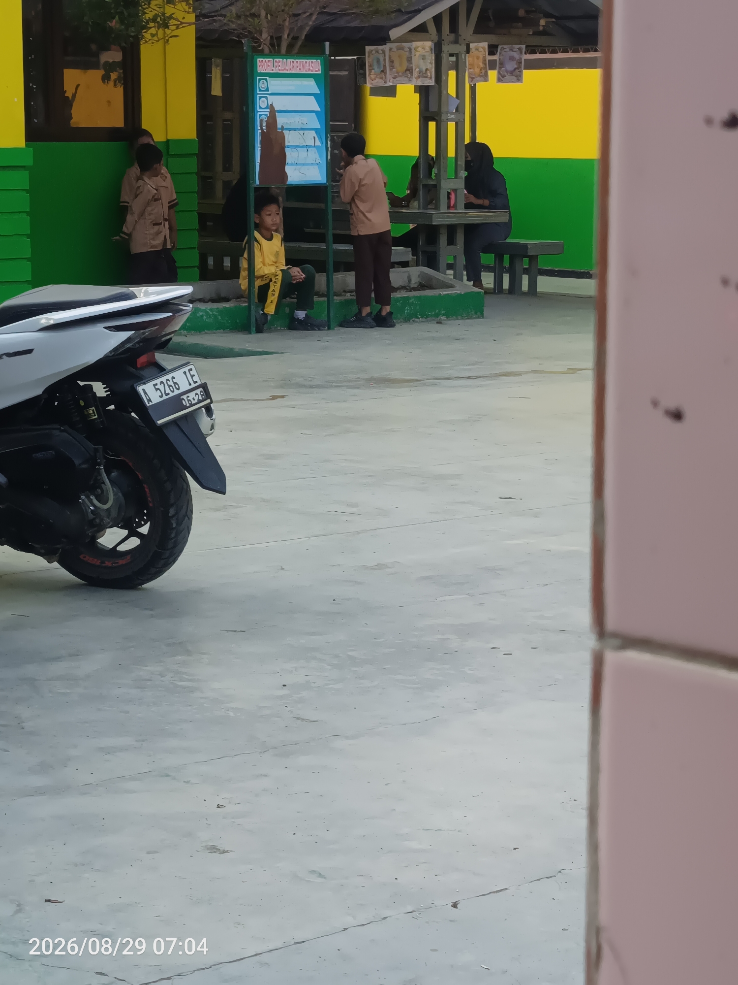 In Sangiang, Indonesia, a group of individuals, likely students, are visible outdoors. In the foreground, the rear of a white motorcycle with a license plate reading "A 5266 IE" is parked on a concrete ground. Behind the motorcycle, a young boy in a yellow shirt and dark pants sits on a green bench next to a blue informational board. Further back, two individuals in tan uniforms stand near a yellow and green building. To the right, two people are seated at a table under a shelter, one wearing a black hijab and the other partially obscured. The environment appears to be a school or public area with a paved ground and a building with brightly colored walls. A timestamp in the bottom left corner indicates the image was captured on "2026/08/29 07:04".
