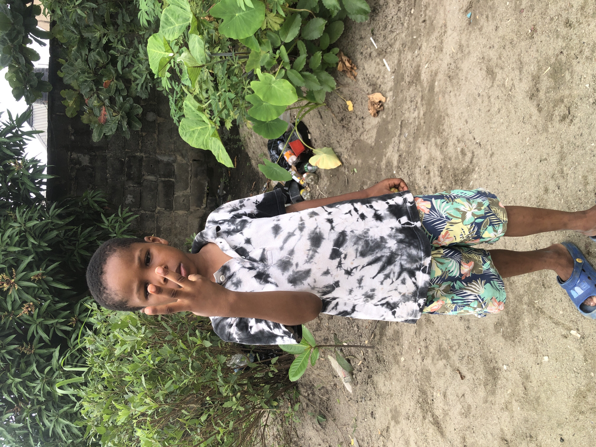 A young Black boy stands facing forward in a slightly dusty outdoor area, likely in Ado, Nigeria. He is wearing a black and white tie-dye polo shirt and multi-colored floral patterned shorts. The boy is holding up three fingers on his right hand, with his thumb extended and middle, ring, and pinky fingers bent. Behind him, lush green foliage and trees form a dense backdrop, with a grey brick wall visible in the upper portion of the image. In the lower right quadrant, a pile of discarded plastic bottles, bags, and a red container is visible on the ground amidst the greenery. The ground is composed of sand and dirt, with scattered dry leaves and small debris. The boy is wearing blue clogs on his feet.