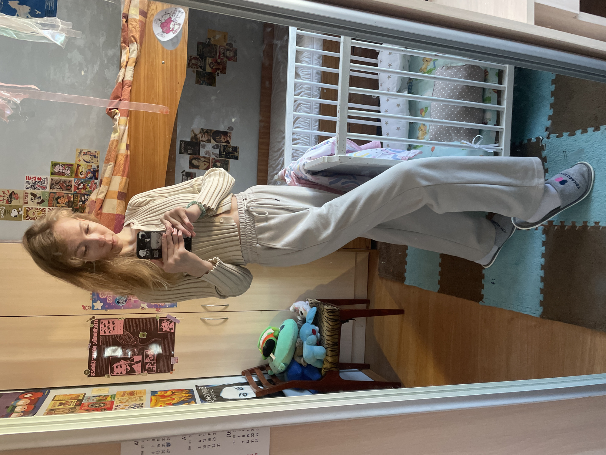 A mirrored selfie reveals a fair-skinned person with shoulder-length blonde hair, dressed in a beige knit top and sweatpants, holding a phone up to their face. They stand in front of a closet door adorned with various stickers and a calendar, beside a small wooden chair stacked with stuffed animals. The mirror reflects a room featuring a crib and a playmat, with a wall covered in decorative images. The reflection suggests the setting is indoors and likely residential. There is no verifiable information on the location within Siva, Russia from the provided image and OCR.