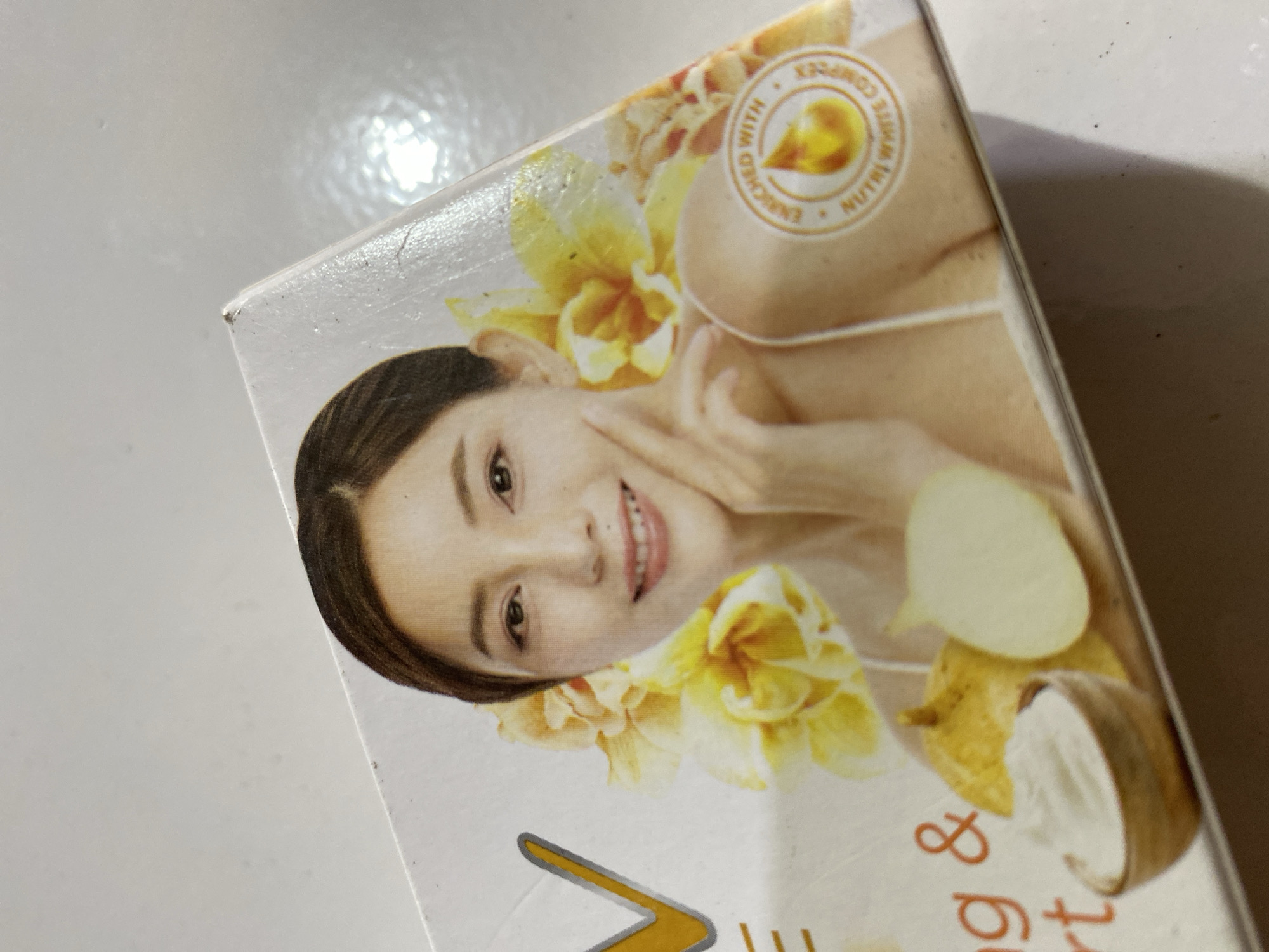 The image shows a close-up of a product box with a woman's face featured on it. The woman is smiling and has her hand near her face. There are yellow flowers behind her. Text on the box is partially visible, including what appears to be the letter "V" and the words "ENRICHED WITH NUTRI WHITE COMPLEX." The box is likely a cosmetic product. The environment appears to be a neutral, indoor setting, possibly a table or countertop, with a slightly reflective surface. There is no information in the image that would suggest a location in Ta', Indonesia.