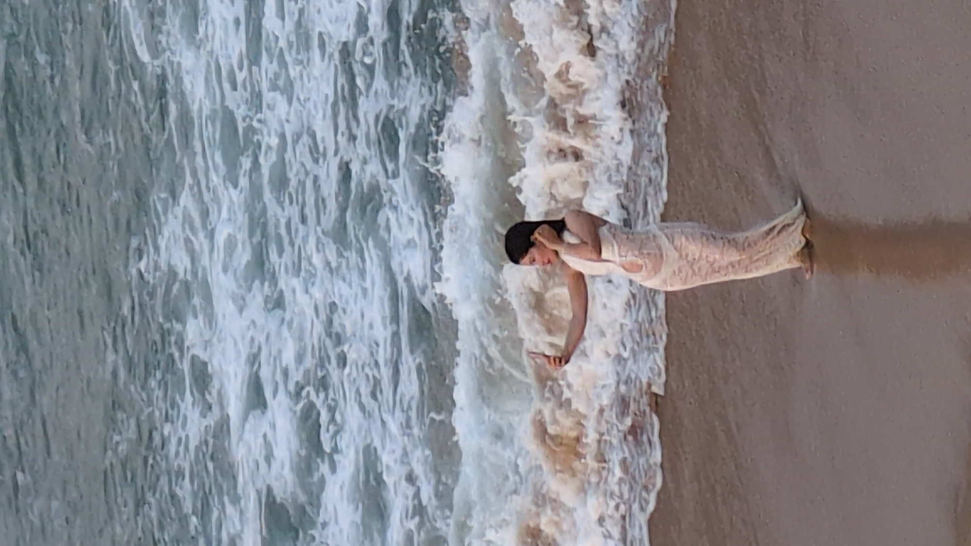 A woman wearing a white, lace-like dress stands at the edge of the beach at Tuy Hòa, Vietnam, in front of the South China Sea. She is looking down at her phone, holding it in her right hand. The ocean waves are turbulent and splashing around her feet. The beach sand is visible to her right. The perspective is from above, looking down at a slight angle.