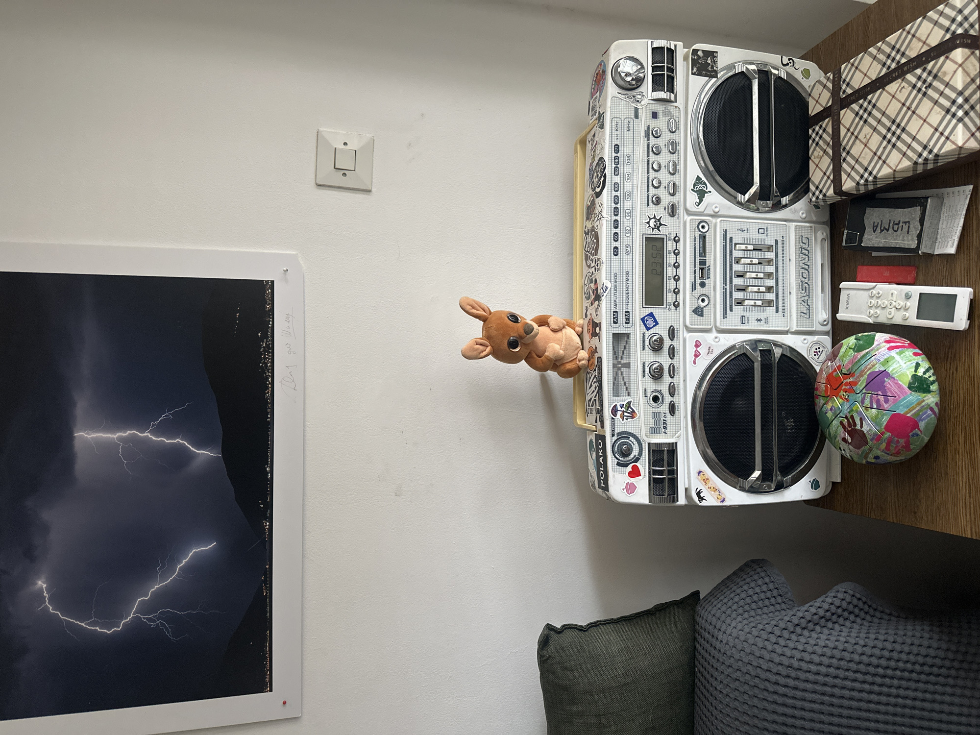 The scene is an interior view within a residential space in Budva, Montenegro, featuring a collection of objects displayed on a wooden surface against a white, textured wall.
On the wall, a large framed photographic print depicts a dramatic nighttime scene with two distinct lightning bolts illuminating a dark, mountainous landscape over a body of water with distant city lights. The print has a white border with a red pushpin in the bottom-left corner and handwritten text on the bottom right. To the right of the print, a white, single-gang electrical light switch is mounted on the wall.
On the far left, a dark green textured cushion is partially visible, with a dark grey waffle-knit blanket draped over an unseen furniture item.
The central focus on the wooden surface is a white LASONIC brand portable stereo (boombox) adorned with numerous stickers, including "POLAKO," a red heart, a mushroom, and various other graphics. Its front panel features two large speaker grilles, an equalizer, and a digital display showing "23:52." Perched on top of the boombox is a small, light brown plush toy resembling a kangaroo or similar marsupial. To the left of the boombox, a spherical object, possibly a small decorative drum, is visible, covered in a colorful, abstract pattern including handprints in multiple colors.
To the right of the boombox, a white remote control labeled "XVAIΛ" lies on the wooden surface. Next to it, a black rectangular case or wallet with "LIAMA" written in white is positioned over a crumpled paper receipt. Furthest to the right, a rectangular gift box wrapped in a brown and tan tartan (plaid) pattern, secured with a ribbon marked "SECRET WISH," rests on the surface, showing minor signs of wear.
No individuals or active interactions are present in the image.
