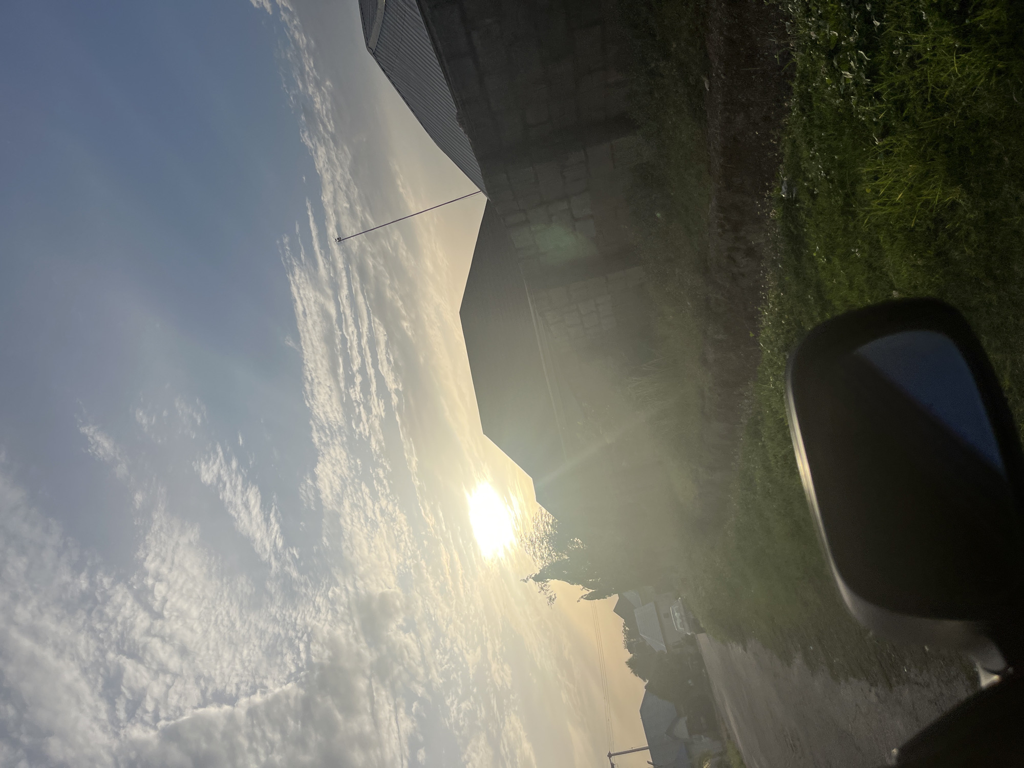 The image depicts a partly cloudy sky at sunrise or sunset. The sun is obscured by clouds, creating a bright glare. A building with a metal roof is visible on the left side of the frame. A grassy area and a road are on the right side of the image. A side-view mirror of a vehicle is visible in the foreground. The location cannot be determined from the image.