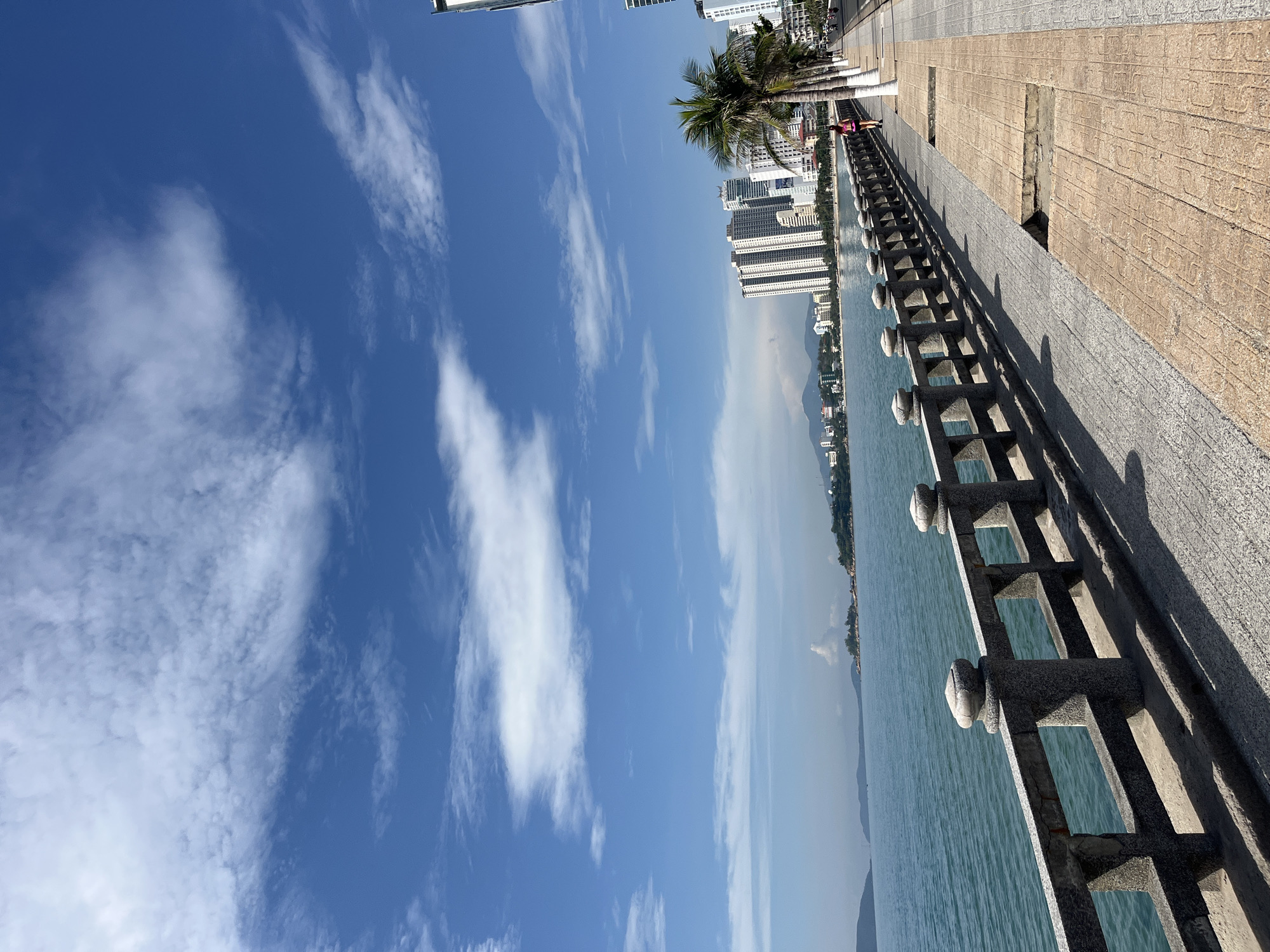 The image shows a seaside view from Bac Nha Trang Ward, Vietnam. A weathered concrete fence runs along the water's edge. The sea is calm with a slight haze. Behind the fence, a tiled walkway extends along the waterfront. Buildings, including high-rise apartments, and palm trees line the walkway. A clear blue sky with scattered white clouds dominates the upper portion of the image. A person is visible walking on the path further down the road.