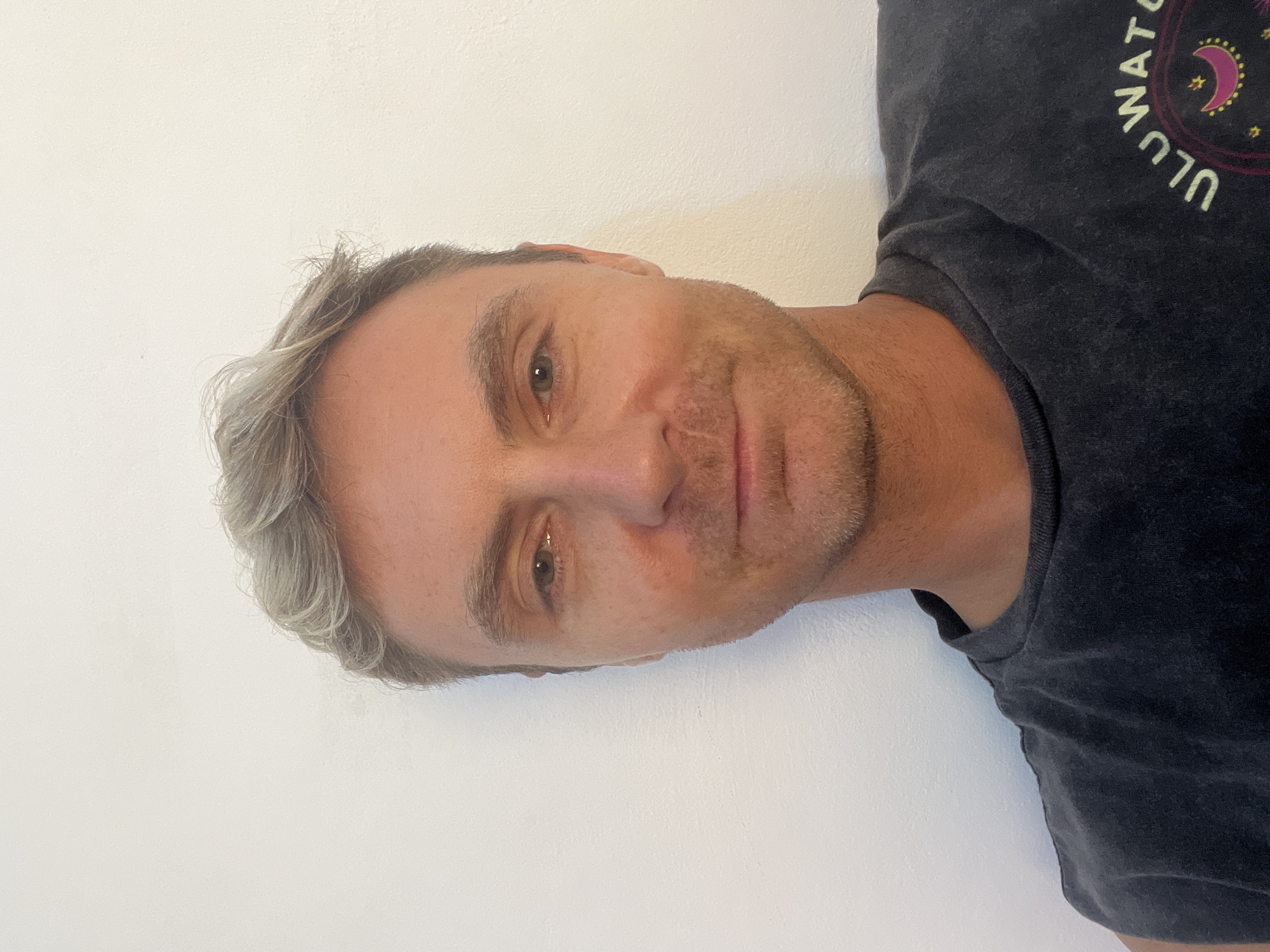 A medium shot of a man against a white wall. He has short, graying hair and light stubble on his face. He is wearing a dark t-shirt with a circular design that includes the word "ULU." The picture was taken in Celuk, Indonesia.