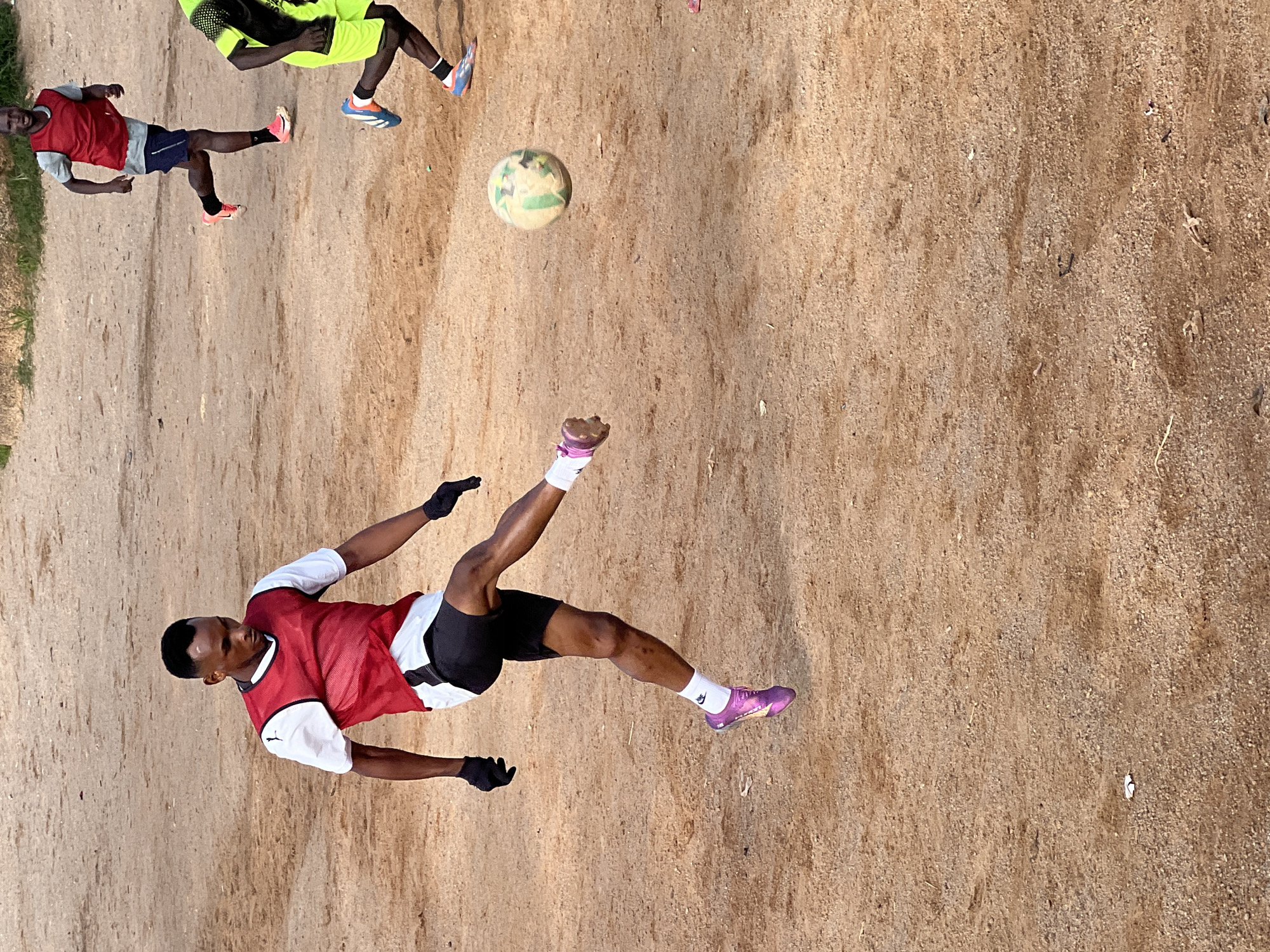 An outdoor daytime full shot captures a young Black male soccer player in mid-kick on a dirt field in Jos, Nigeria. He is wearing a white t-shirt with a red bib, black shorts, black gloves, white socks, and purple soccer cleats. His left leg is extended forward, kicking a white and green soccer ball that is airborne to his right. His body is angled towards the right, with his head down, focused on the ball. In the background, two other male soccer players are visible, one in a red bib and dark shorts, and another in a bright green and black soccer jersey and shorts. The ground is composed of dry, brown dirt and gravel, with sparse vegetation visible in the distance. The lighting suggests a sunny day.
