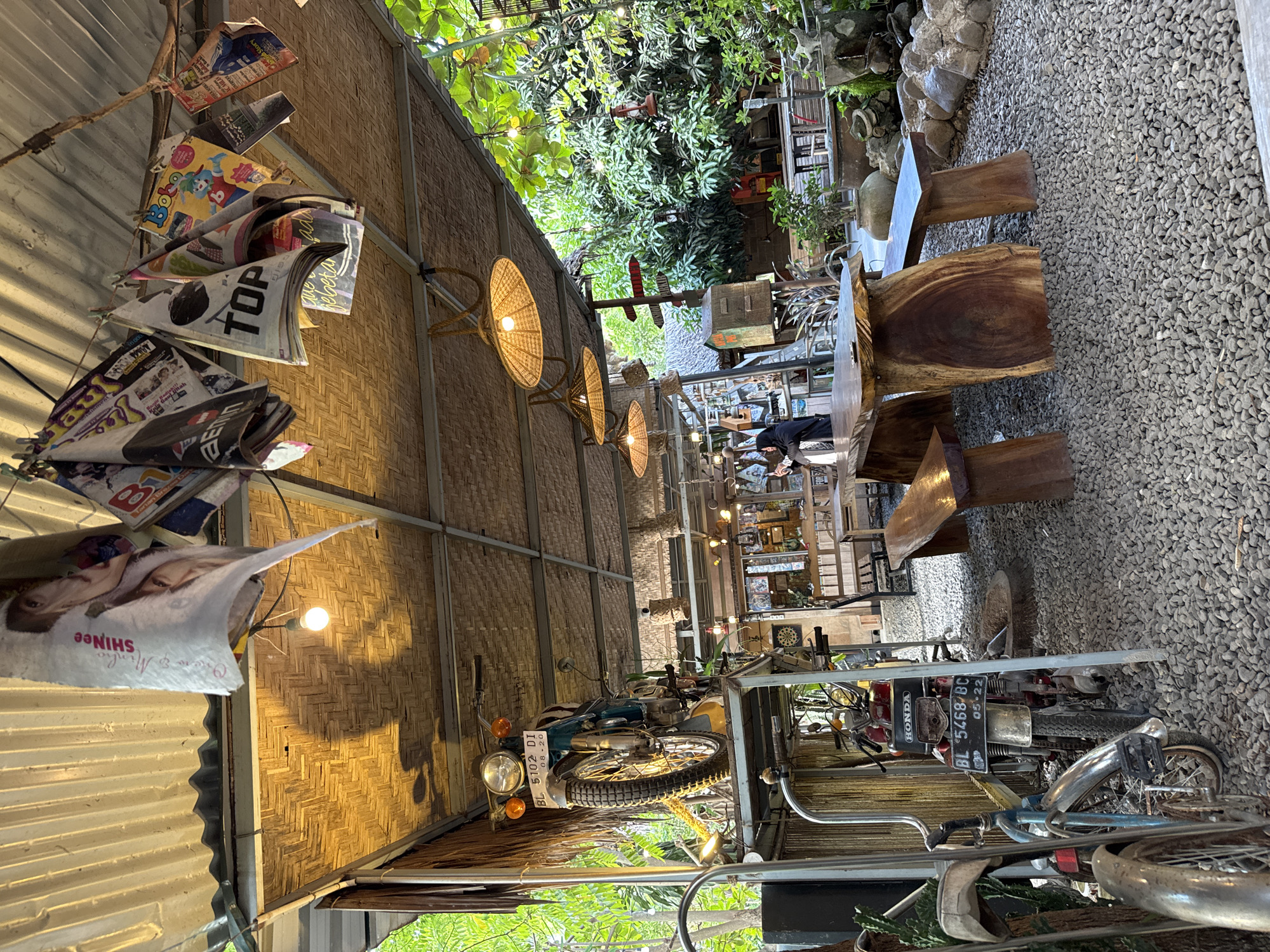 This image depicts an outdoor seating area of a cafe in Cotbadatunong, Indonesia, characterized by a rustic and eclectic ambiance. The roof structure is a corrugated metal sheet overlaying a woven bamboo ceiling, with several hanging lamps featuring straw shades. Magazines and papers are clipped to a wire suspended from the roof, displaying various graphics and text, including one with "SHINEE" and another with "TOP".
Dominating the left side of the scene is a vintage motorcycle, mounted on a platform supported by metal scaffolding. Below this, another motorcycle is visible, leaning against a vertical support. Both motorcycles appear to be older models, contributing to the cafe's vintage aesthetic.
The seating area is furnished with a large, thick wooden table supported by two substantial wooden legs, and matching wooden benches. The ground is covered in grey gravel. In the background, more seating arrangements are visible, along with greenery from trees and plants, suggesting a garden or outdoor setting. A few decorative elements, such as lanterns and what appears to be a birdcage, are also present. A person, wearing a hijab and dark clothing, is partially visible in the background, interacting with something at a table. The overall impression is a casual, open-air establishment with a bohemian or retro vibe.