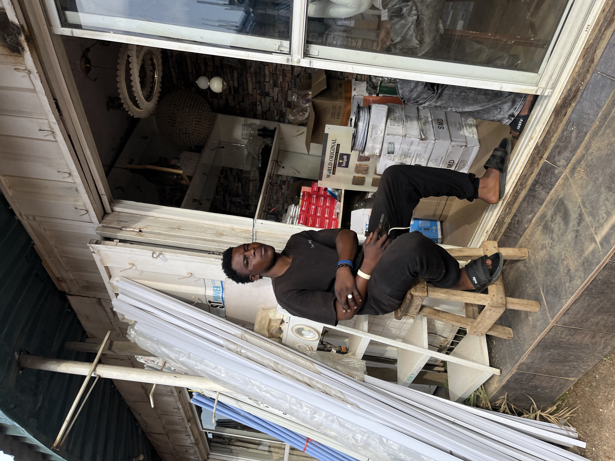 This media file shows a young Black man sitting on a wooden stool in front of a store or workshop. He is wearing a dark t-shirt, black pants, and black sandals. He has a ring on his right hand and bracelets on his wrists. The man is looking towards the camera with a neutral expression.
The setting appears to be an outdoor market or a workshop, with shelves filled with various items behind the man. Some visible items include boxes with text like "SMD," "MD," and "WORLD ORIGINAL." There are also what appear to be building materials or fixtures stacked in front of the store. The time of day seems to be daytime, possibly late morning or afternoon, given the lighting. The overall impression is one of a busy, commercial environment.
The text visible in the image includes brand names like "Nike," "LUOFU," and "Fumigalli," as well as descriptions such as "Switches & Sockets Italian Standard," and "MADE IN CHINA." The word "STPOMER" is also visible. The location context indicates this scene is in Gura Top, Nigeria.
