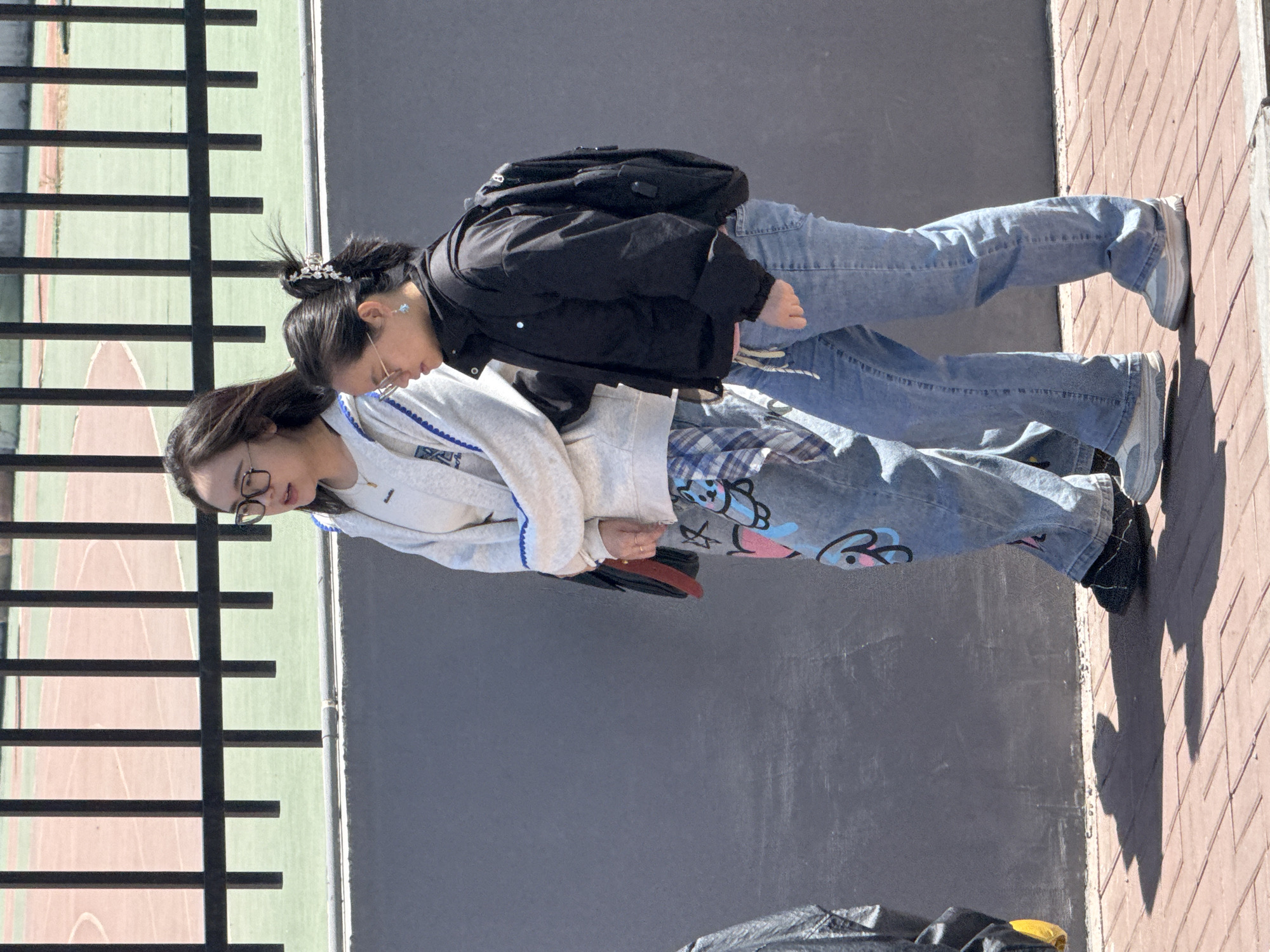 Two young women are standing outdoors. The woman closest to the camera is wearing glasses, a light-colored sweatshirt with blue trim, and patterned light-blue jeans, and black shoes. She is holding a reddish-brown clutch purse. The woman further from the camera is wearing glasses, a black jacket, light-blue jeans, and grey shoes. She has a black backpack on. They are standing near a low, black metal fence. Behind the fence is a tennis court, and next to them is a grey wall and red bricks. Zhongtun, China cannot be confirmed.