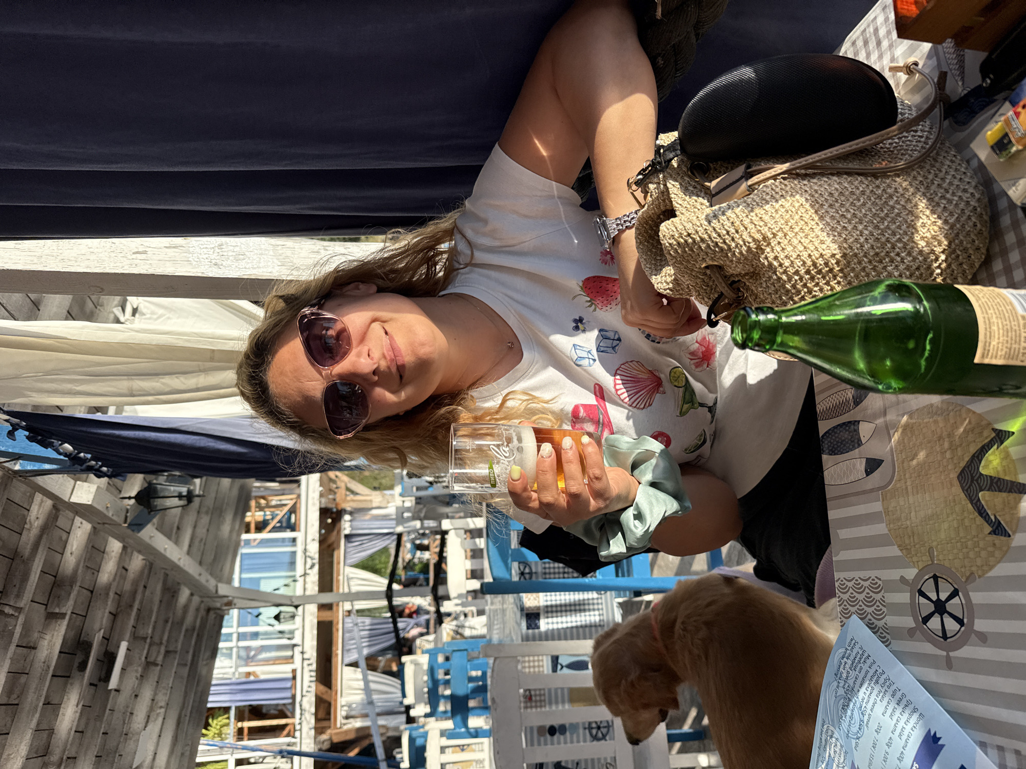 A medium shot captures a young blonde woman with sunglasses and a t-shirt adorned with fruit graphics, holding a glass of beer. She is seated at a table, with a straw bag and a green bottle of beer in front of her. A golden retriever sits to her left, looking away. The scene is set outdoors at a cafe with white and blue chairs, wooden beams overhead, and draped curtains. The overall lighting suggests it is late afternoon or early evening.