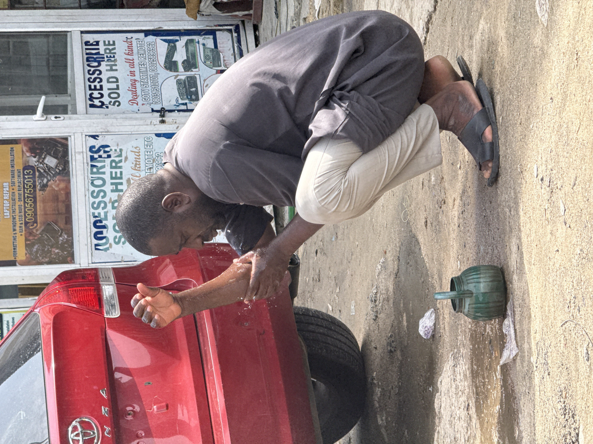 A man in a grey tunic and white pants washes his hands over the rear of a red Toyota SUV. He kneels on the ground, wearing a single black sandal. Next to him is a green watering can. Behind the car is a storefront with signs reading "LAPTOP REPAIR," "ACCESSORIES SOLD HERE," and phone numbers. The location is likely in Lagos, Nigeria, as inferred from the phone numbers and general environment.