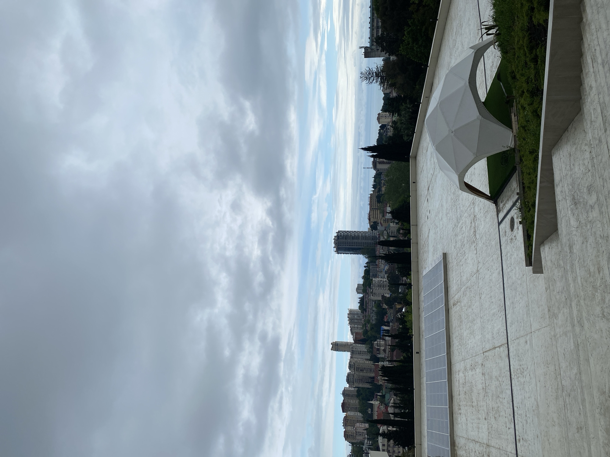 The image shows a view from a rooftop in Sochi, Russia. In the distance is a cityscape with buildings of varying heights, interspersed with trees and vegetation. The sky is overcast with a mix of white and gray clouds. On the rooftop, there is a white geometric structure next to a strip of green vegetation and a vertical row of solar panels attached to the surface of the building. The rooftop is made of a light-colored material with a textured surface.