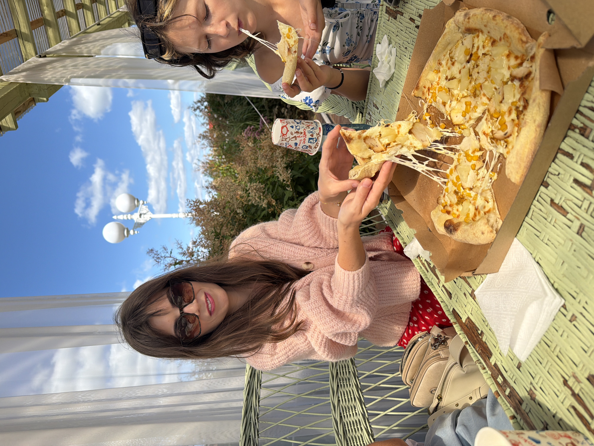 Two young women are dining outdoors at a table under a pergola. The woman on the left, wearing a pink sweater and red polka-dot skirt, is reaching for a slice of pizza from a box on the table. She is wearing sunglasses. The woman on the right, wearing a floral top, is eating a slice of pizza. Both women are seated on a green wicker bench. In the background, there is a blue sky with white clouds, trees, and a lamppost. The table is covered with a green woven placemat. There are also paper cups and napkins on the table. The location is identified as Severynivka, Ukraine.