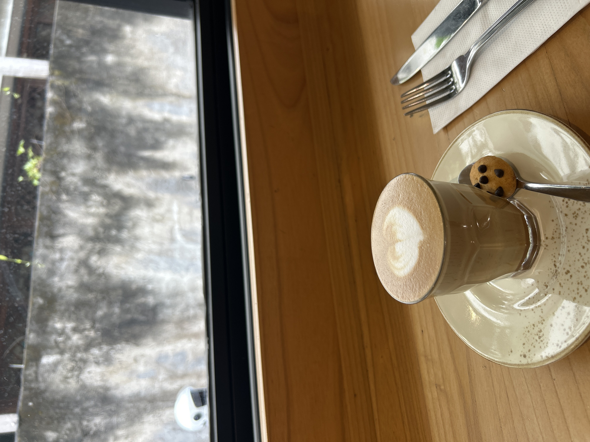 A top-down, high-angle shot presents a cafe scene in Kuta Selatan, Indonesia. To the left, a window with a black frame reveals a blurry, indistinct view of the outside. Next to it, a wooden table holds a glass cup of latte with latte art, sitting on a saucer. A small chocolate chip cookie rests on a spoon. A white napkin with silverware (a fork and knife) is placed on the upper right. The overall impression is of a cafe setting in Indonesia.