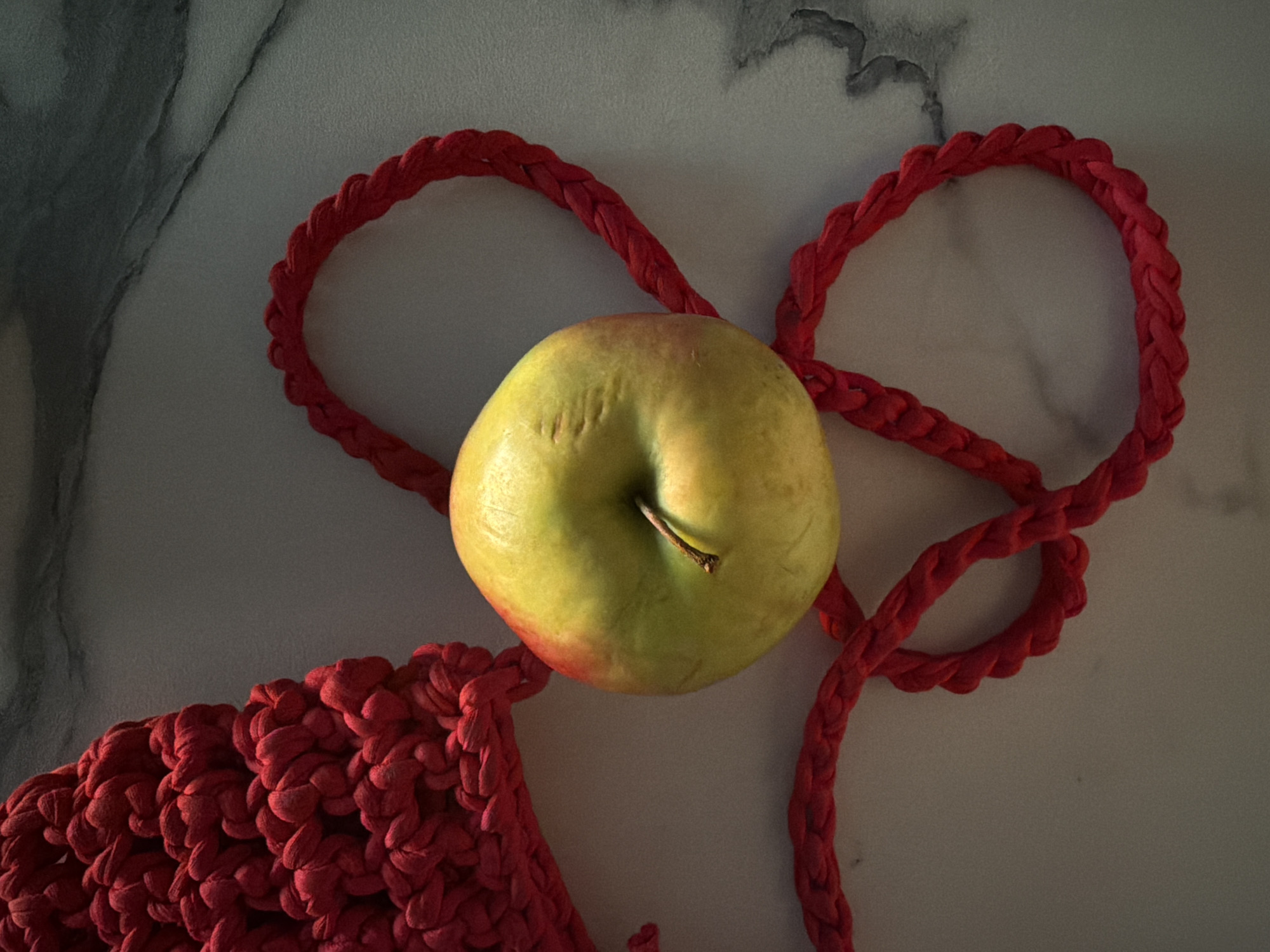 A top-down shot captures a yellow-green apple resting on a section of a red, braided textile. The braided material forms loops and part of what appears to be a bag structure at the bottom left. The surface beneath the apple and textile has a light gray color with subtle marble-like veining patterns.