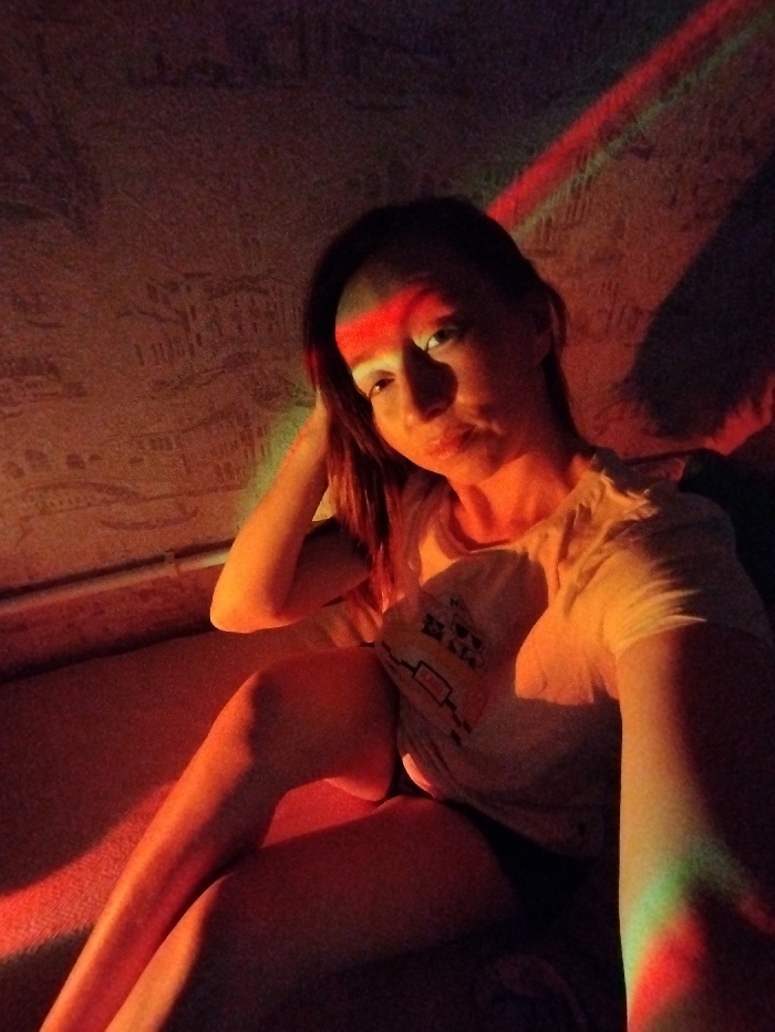 A woman with auburn hair is illuminated by red and green light, casting harsh shadows on her face and surroundings. She wears a light-colored T-shirt with a graphic on it and is seated, with one leg visible. The background appears to be a wall with textured patterns or possibly wallpaper. The lighting creates a vibrant and dramatic effect, obscuring some details of the scene.