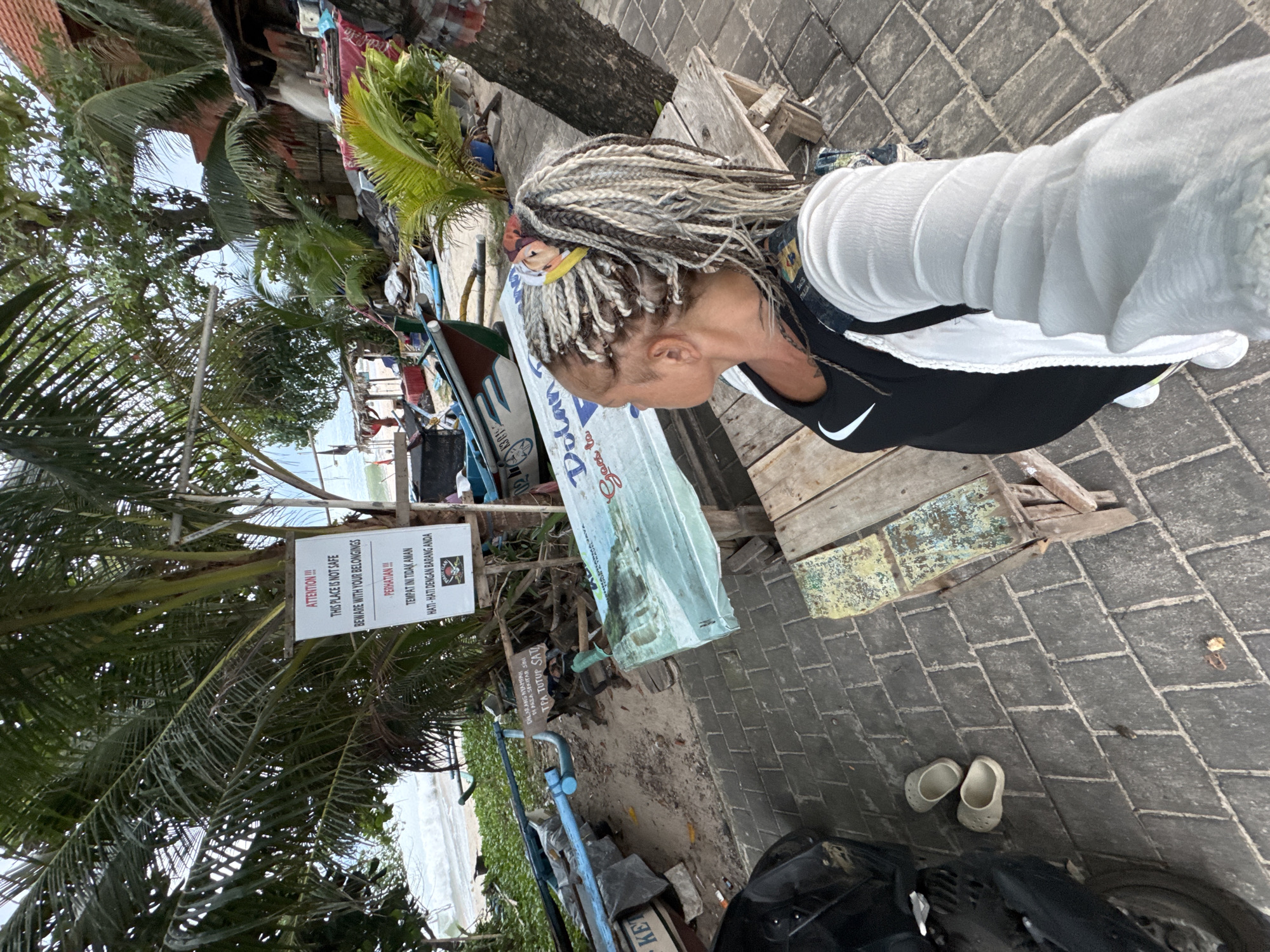 The image, taken in Kuta, Indonesia, shows a woman with braids standing on a brick walkway. A motorcycle is parked nearby, and a pair of white shoes rests on the bricks. In the background, there are palm trees, a boat, and a sign warning people to beware of their belongings because the area is not safe. The beach and ocean are visible in the distance.