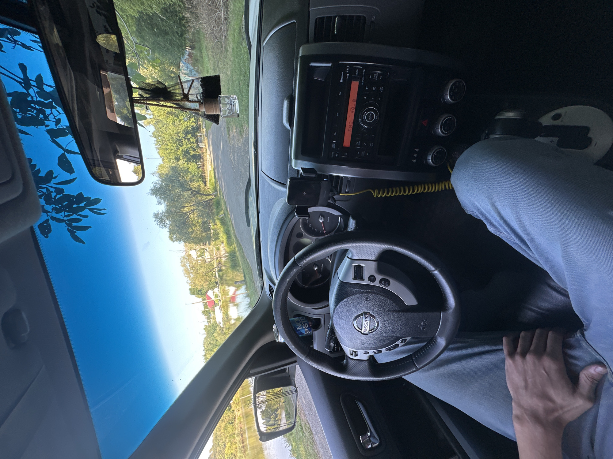 The image shows the interior of a Nissan vehicle, viewed from the driver's perspective. The driver's legs, clad in blue jeans, are visible, with one hand resting on their lap. The steering wheel, with the Nissan logo, is in the foreground. The car's dashboard includes a radio display showing "FM2 90.0", a mobile phone mount with a phone attached, and climate control knobs. A yellow coiled cable runs from the mount. A car air freshener hangs from the rearview mirror. Outside the car, visible through the windshield and side windows, is a body of water, possibly a lake, bordered by trees and a road. In the distance, red-roofed buildings can be seen on the opposite shore of the water. The overall setting appears to be a rural or semi-rural area, potentially near Makiv, Ukraine, given the context. The lighting suggests it is daytime, with sunlight illuminating the scene.