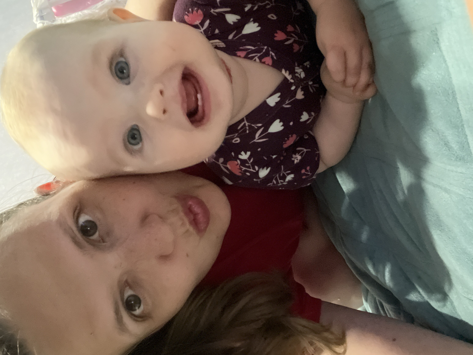 A photo shows a baby and an adult looking at the camera. The baby has blonde hair and blue eyes and is wearing a purple floral shirt. The adult has brown hair and eyes and is wearing a red shirt. They appear to be lying down on a turquoise blanket. No information suggests this photo was taken in Chaikovsky, Russia.