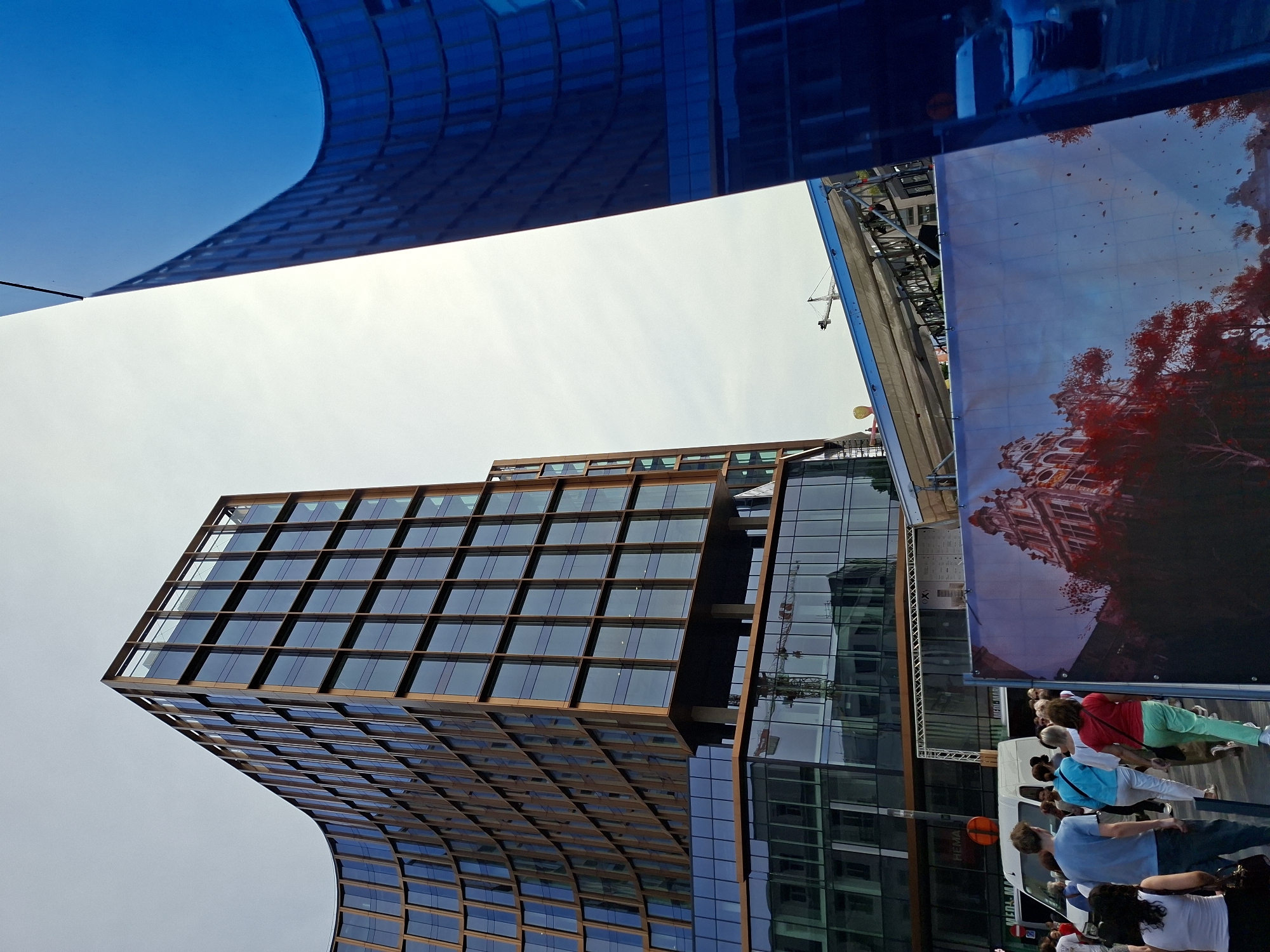 The photograph captures a view of a modern, curved building with a facade of glass and wood, set against a clear sky. In the foreground, a group of people are walking past a large banner depicting an ornate building and red trees. A white van is also visible. To the right, another building with a highly reflective blue facade curves into the frame, mirroring the sky and adjacent structures. The scene appears to be in an urban environment, possibly a city square or street, with construction elements like scaffolding and a crane visible in the mid-ground, suggesting ongoing development. The architectural style of the main building is contemporary, with clean lines and a grid-like pattern of windows framed by wooden elements. The banner in the foreground adds a contrasting element of historical or artistic imagery to the modern urban landscape. The presence of people suggests it's a public space. The overall impression is one of a dynamic, developing urban area. This specific building, characterized by its distinctive curved facade and glass-and-wood exterior, is located at Place de la Gare du Nord in Brussels, Belgium.