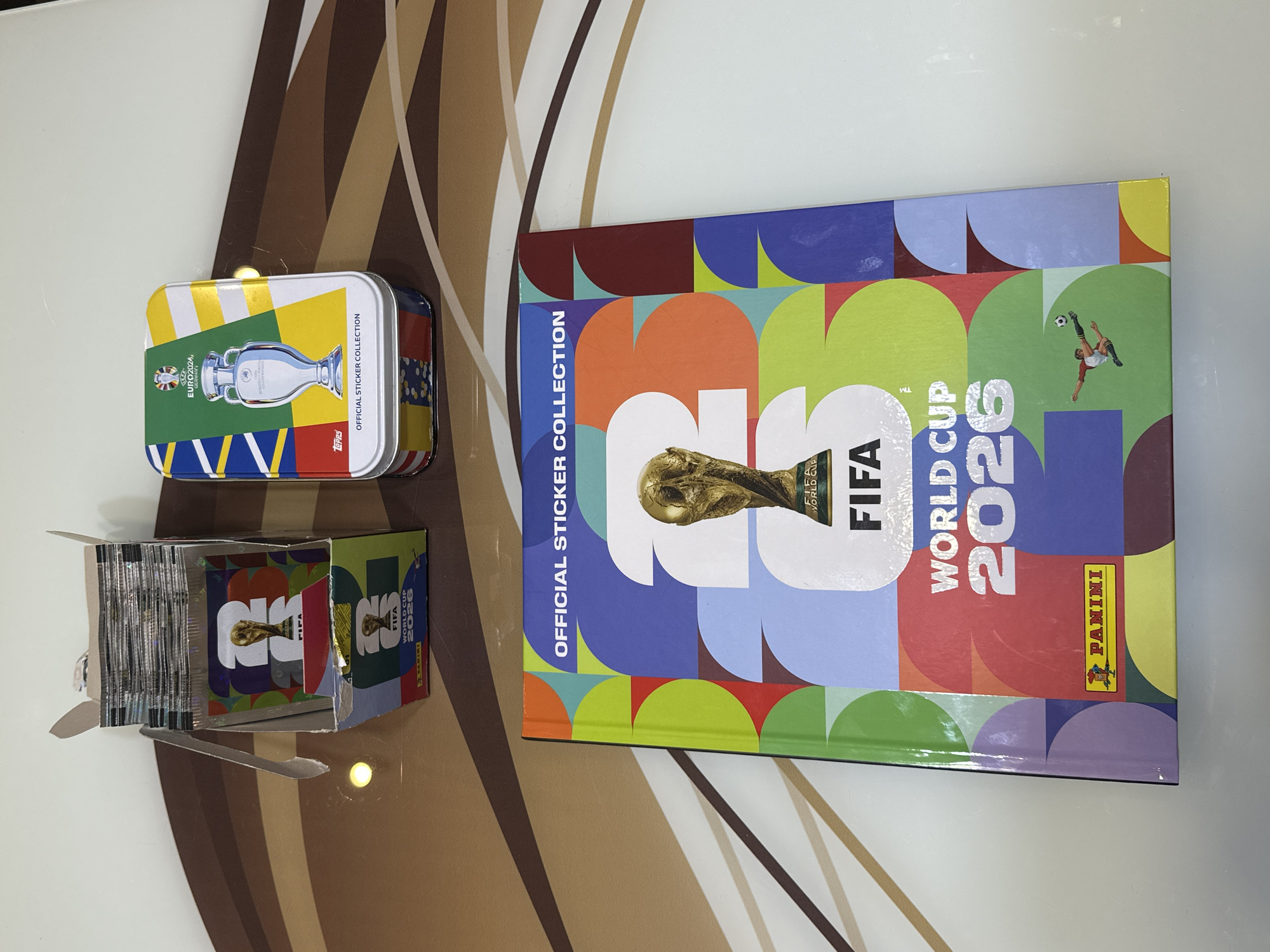 A collection of football sticker memorabilia is displayed on a patterned table in Prisovo, Bulgaria. The primary object is a large, hardcover sticker album titled "OFFICIAL STICKER COLLECTION FIFA WORLD CUP 2026" by Panini. Its cover is vibrant with a mosaic of colorful shapes and features a gold image of the World Cup trophy and a small illustration of a football player in mid-air.
To the left of the album, an open cardboard box holds numerous sealed sticker packets, also branded "FIFA WORLD CUP 2026" and Panini. Further to the left, a rectangular metal tin is visible. This tin is an "OFFICIAL STICKER COLLECTION" for "EURO2024 GERMANY" by Topps, depicting the Euro trophy against a background of national flag colors.
The objects are arranged on a table with a distinctive pattern of broad, curved stripes in shades of brown, beige, and white. The scene is indoors, under what appears to be artificial lighting, causing some reflections on the glossy surfaces of the album and tin. The items suggest an activity of collecting football stickers.